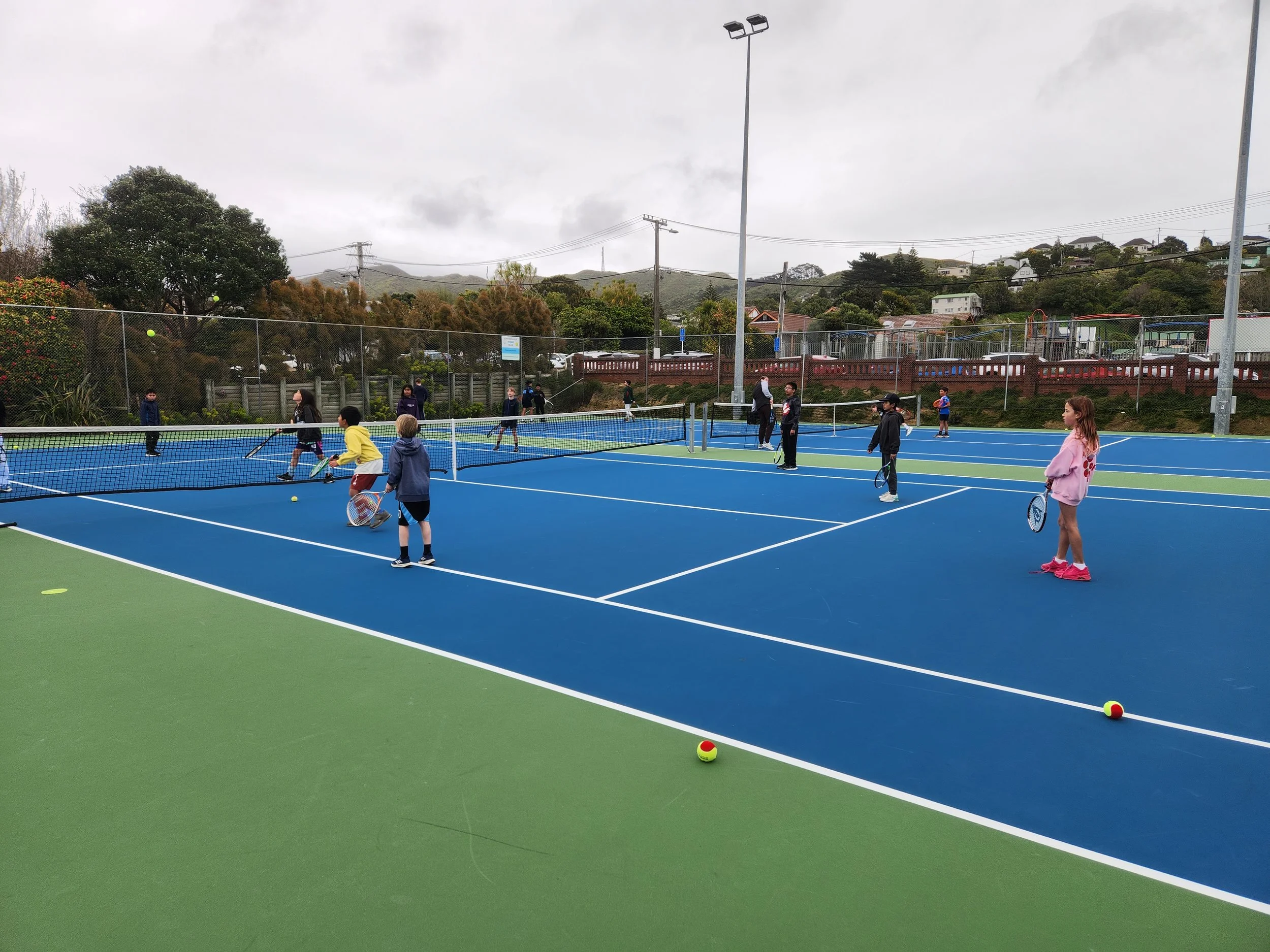 Children and adults playing pickleball on outdoor courts on a cloudy day.