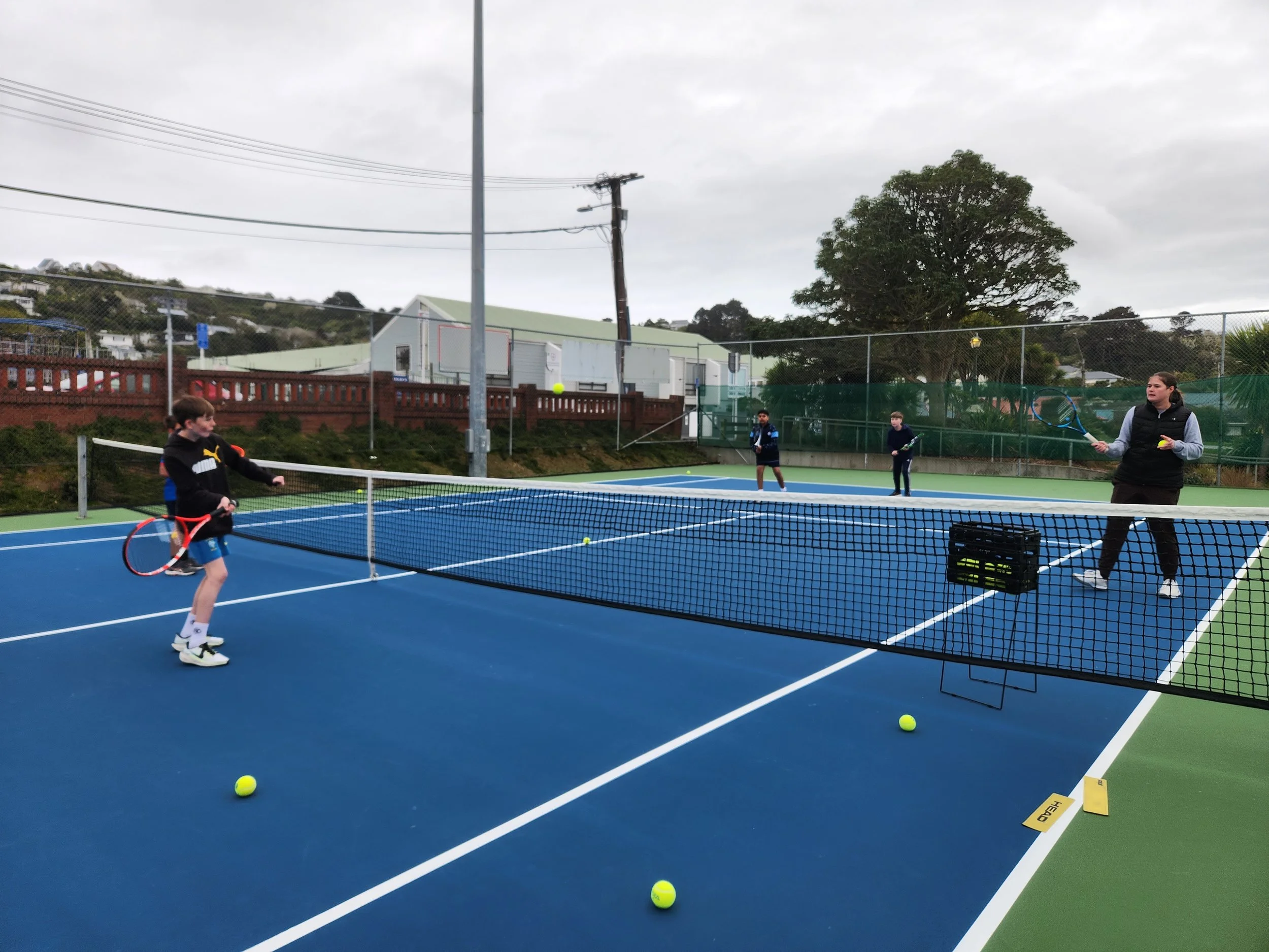 Children and an adult playing tennis on an outdoor court, with tennis balls and a net, on a cloudy day.