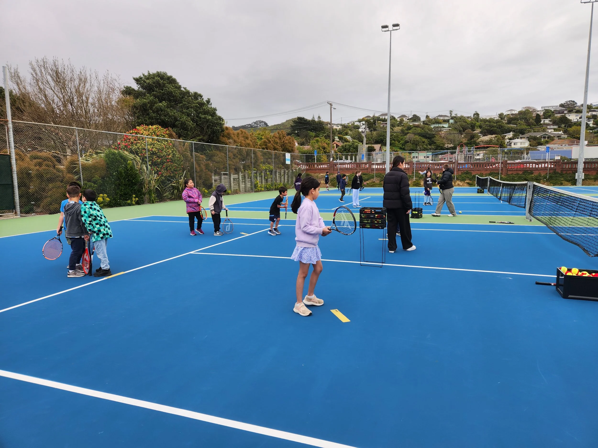 Children and adults playing tennis on outdoor courts on a cloudy day.