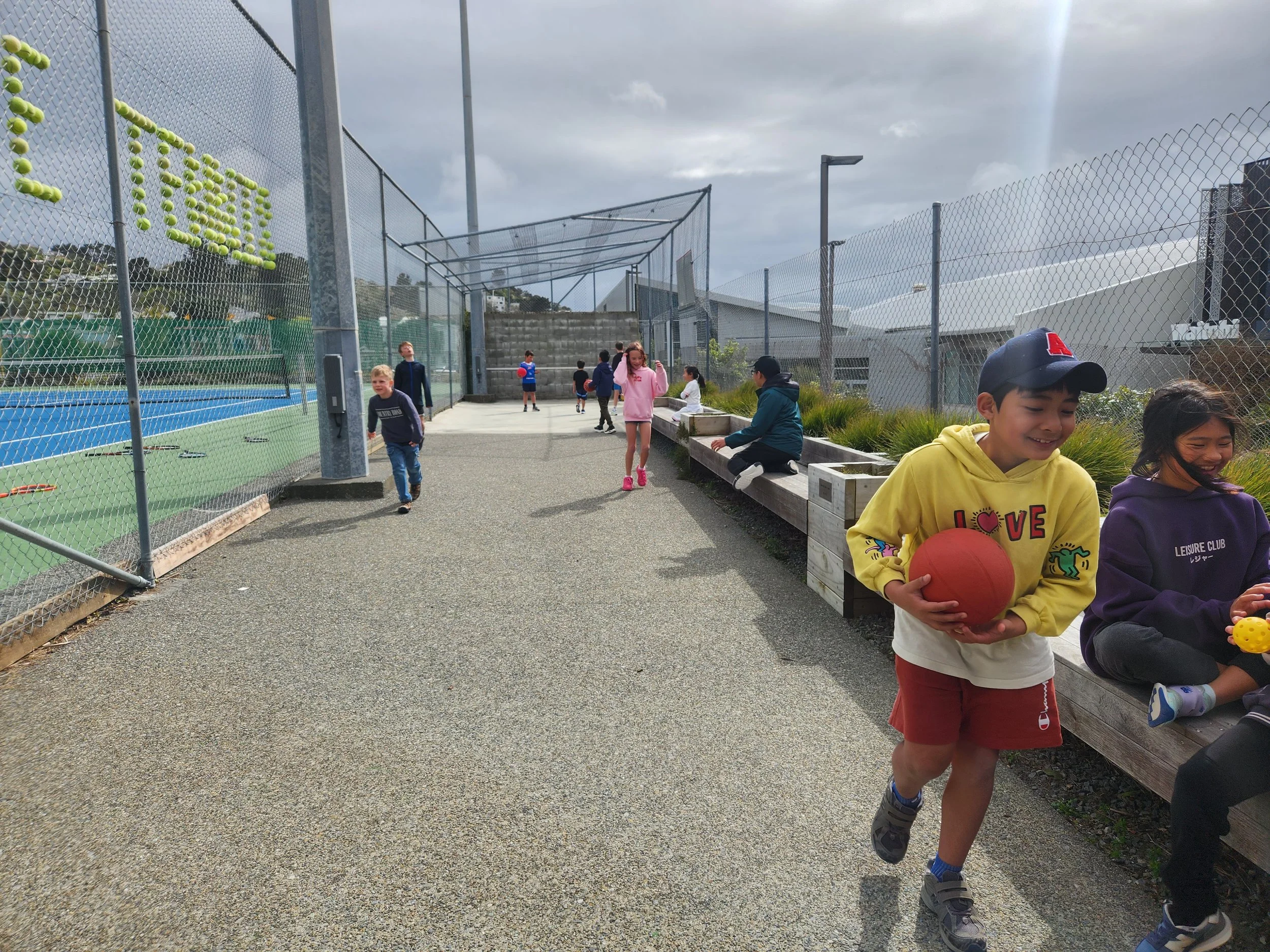 Children playing and sitting near a tennis court and on a bench outdoors on a cloudy day.