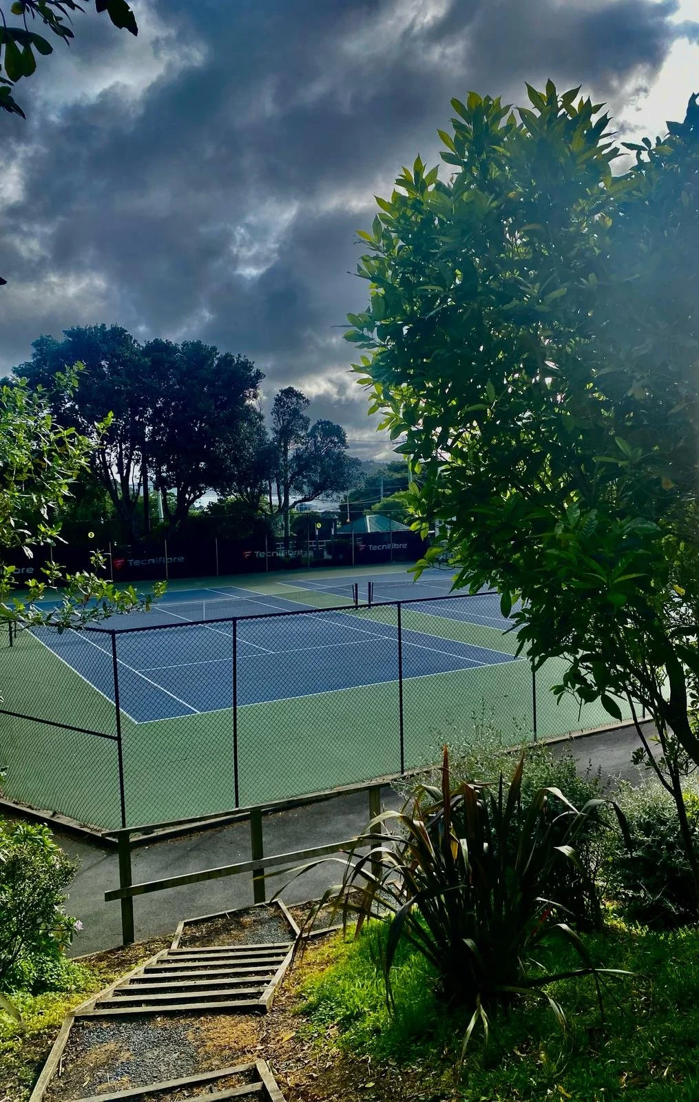 An outdoor tennis court surrounded by greenery and trees under a partly cloudy sky.