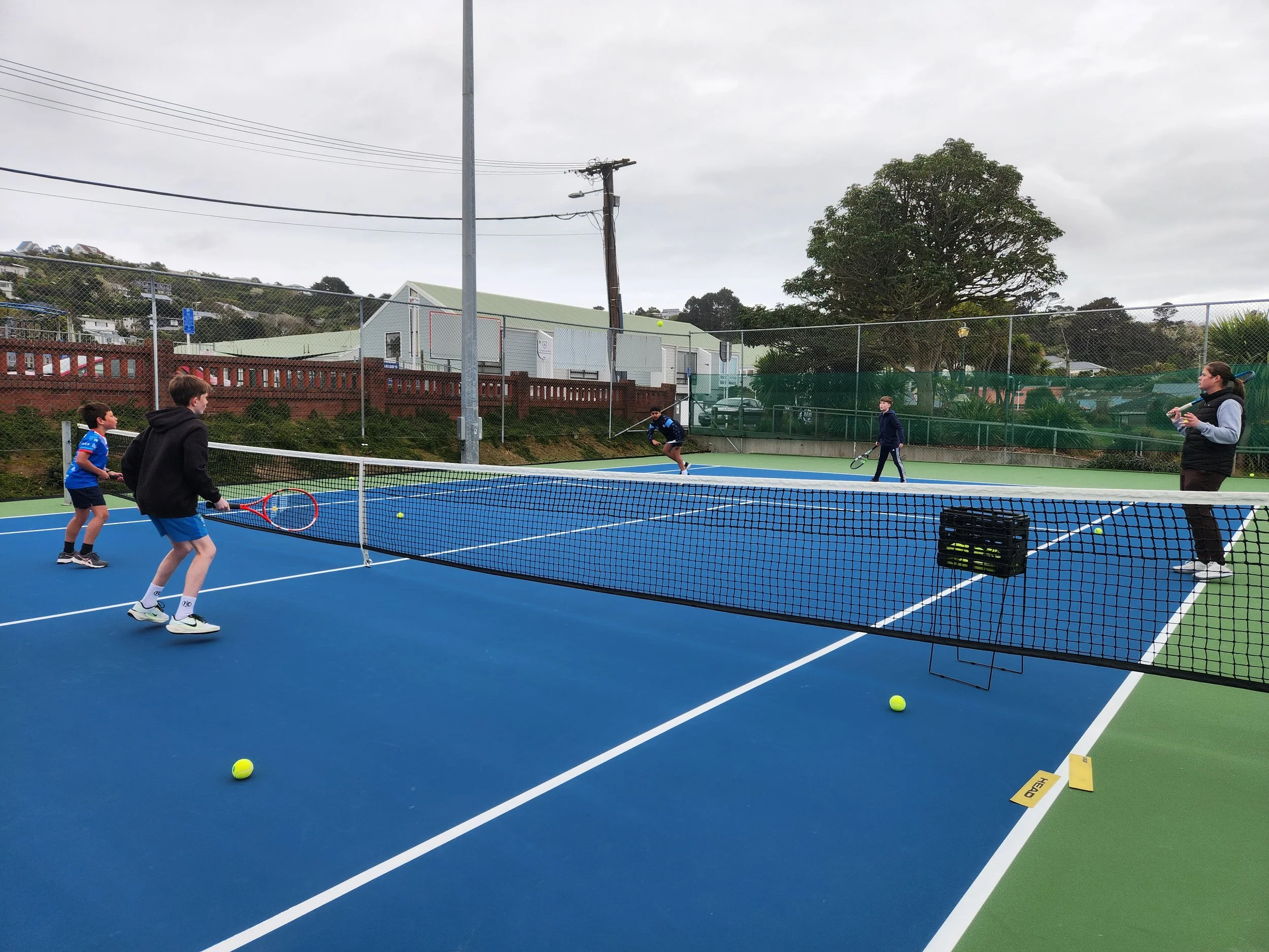 Children and a woman playing tennis on a blue outdoor court, with a net, tennis balls, and a gray sky.