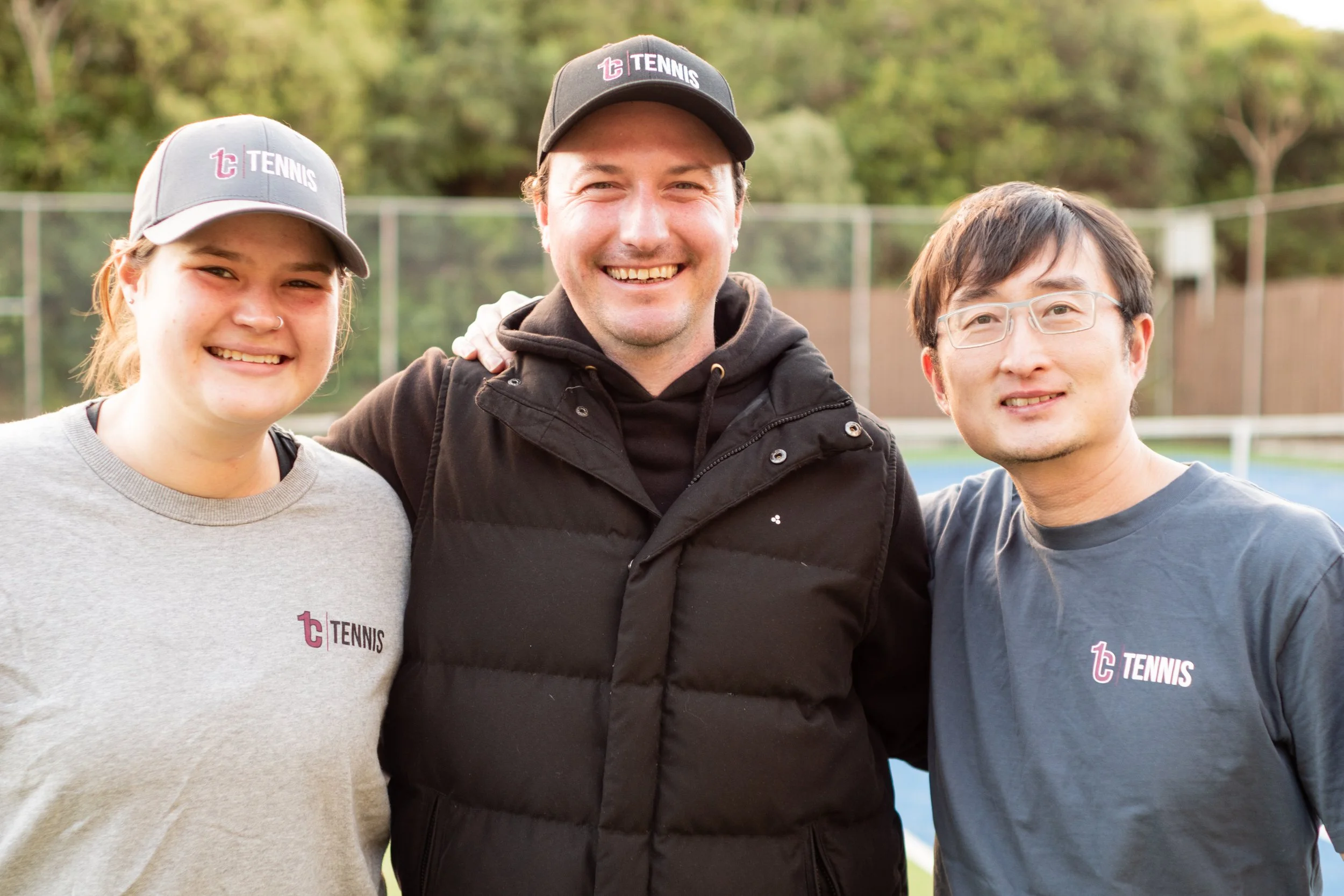 Three people smiling outdoors near a tennis court, two wearing tennis shirts and hats, one in a black jacket.