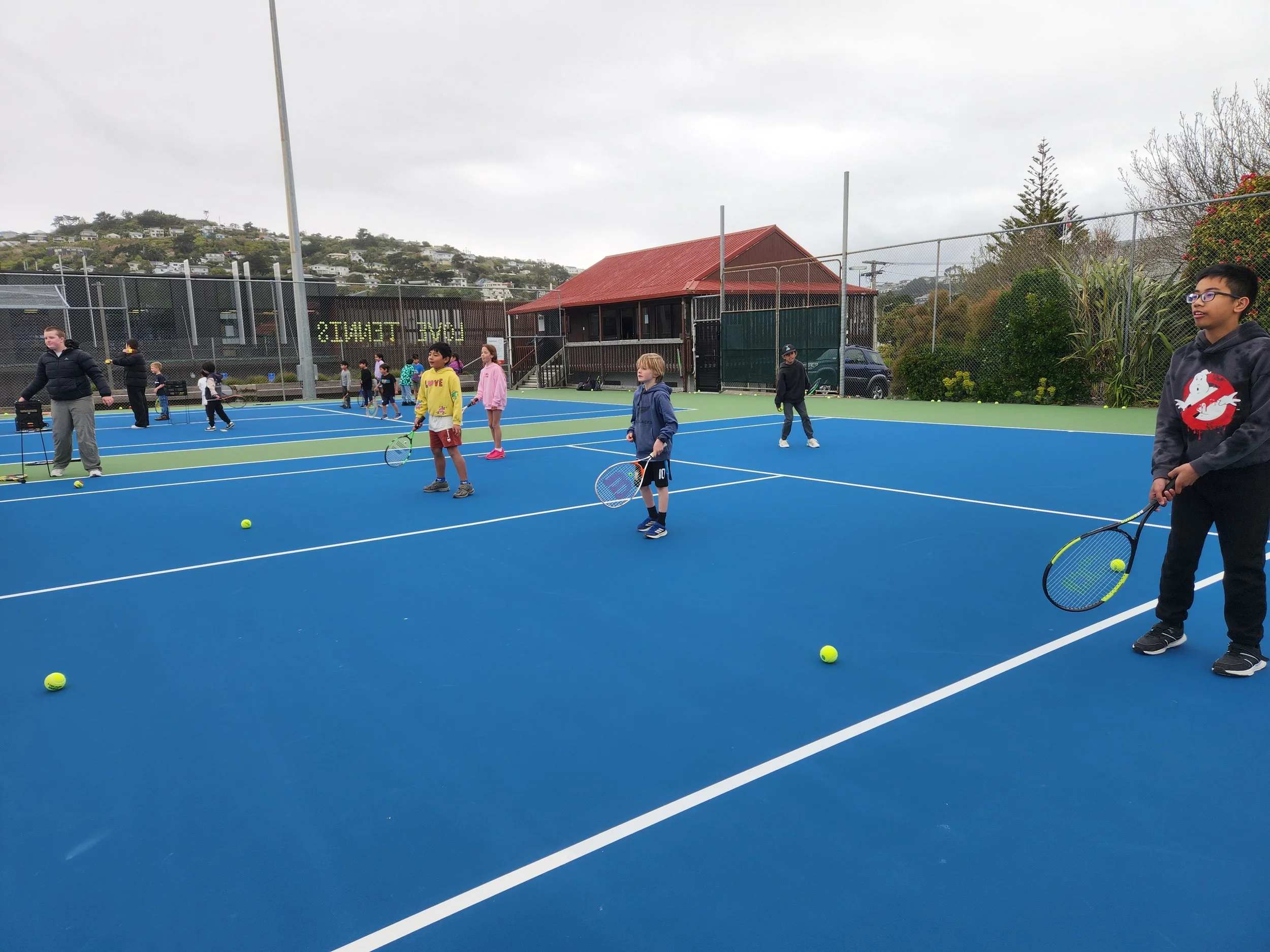 Children playing tennis on a blue outdoor tennis court with a coach, holding tennis rackets and tennis balls, under an overcast sky.