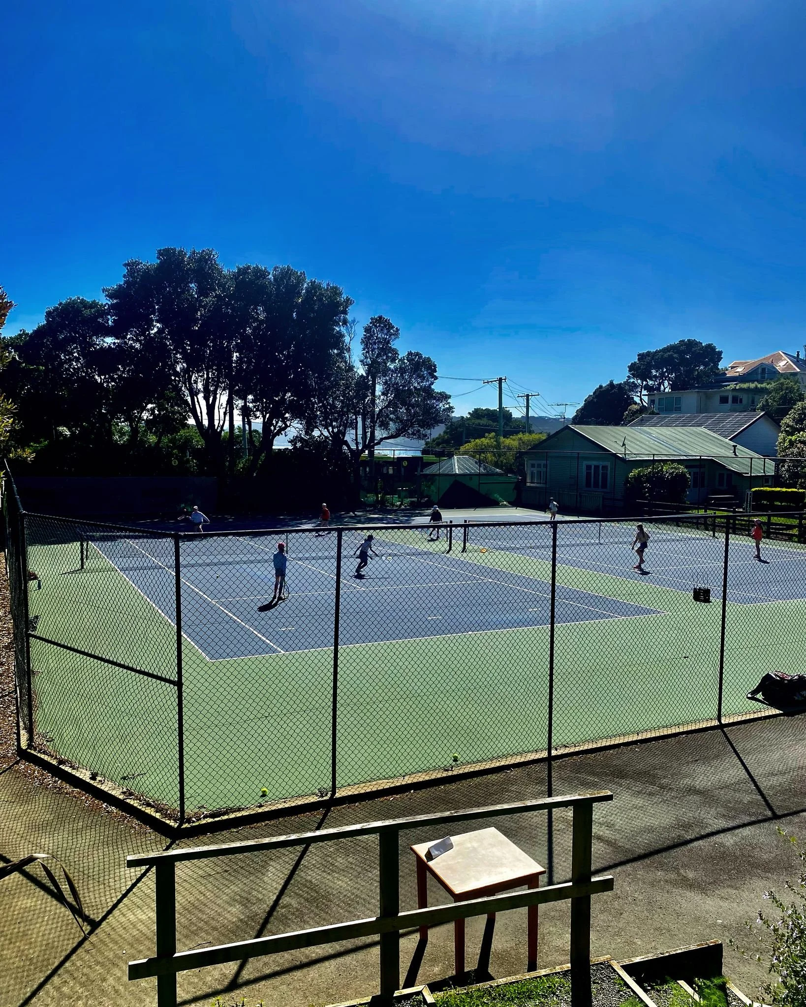 A group of people playing tennis on an outdoor court under a clear blue sky, surrounded by trees and houses.