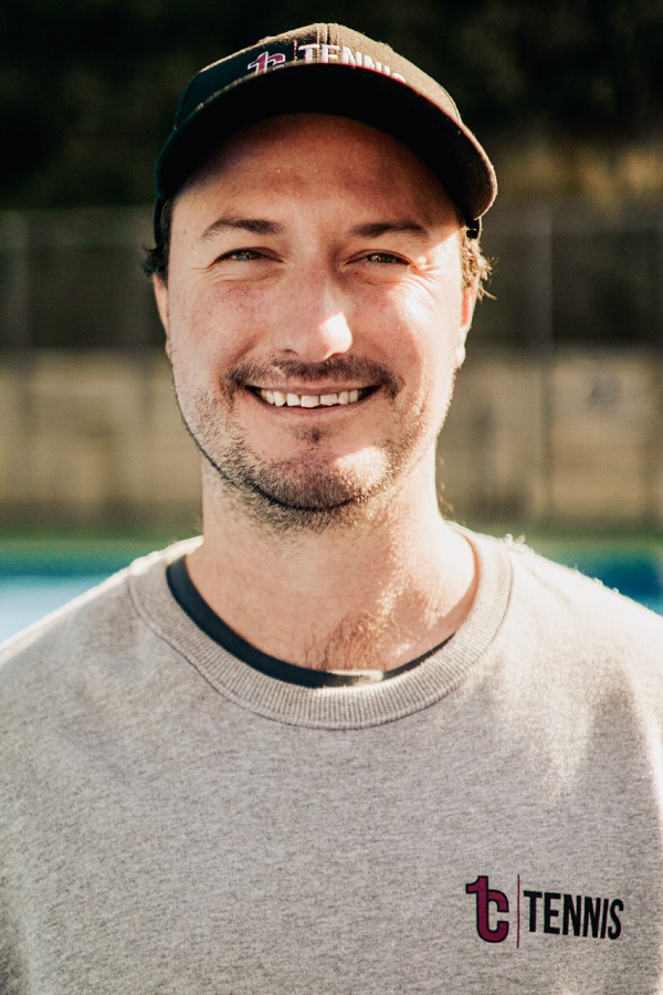 A man smiling outdoors wearing a gray sweatshirt with a tennis logo and a baseball cap.
