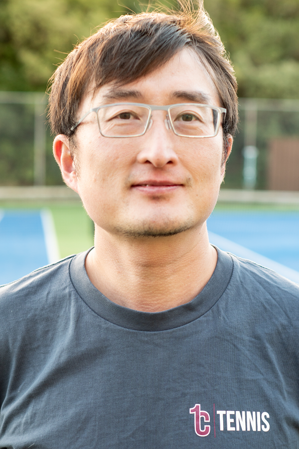 A man wearing glasses and a gray T-shirt with a tennis logo, standing on a tennis court.