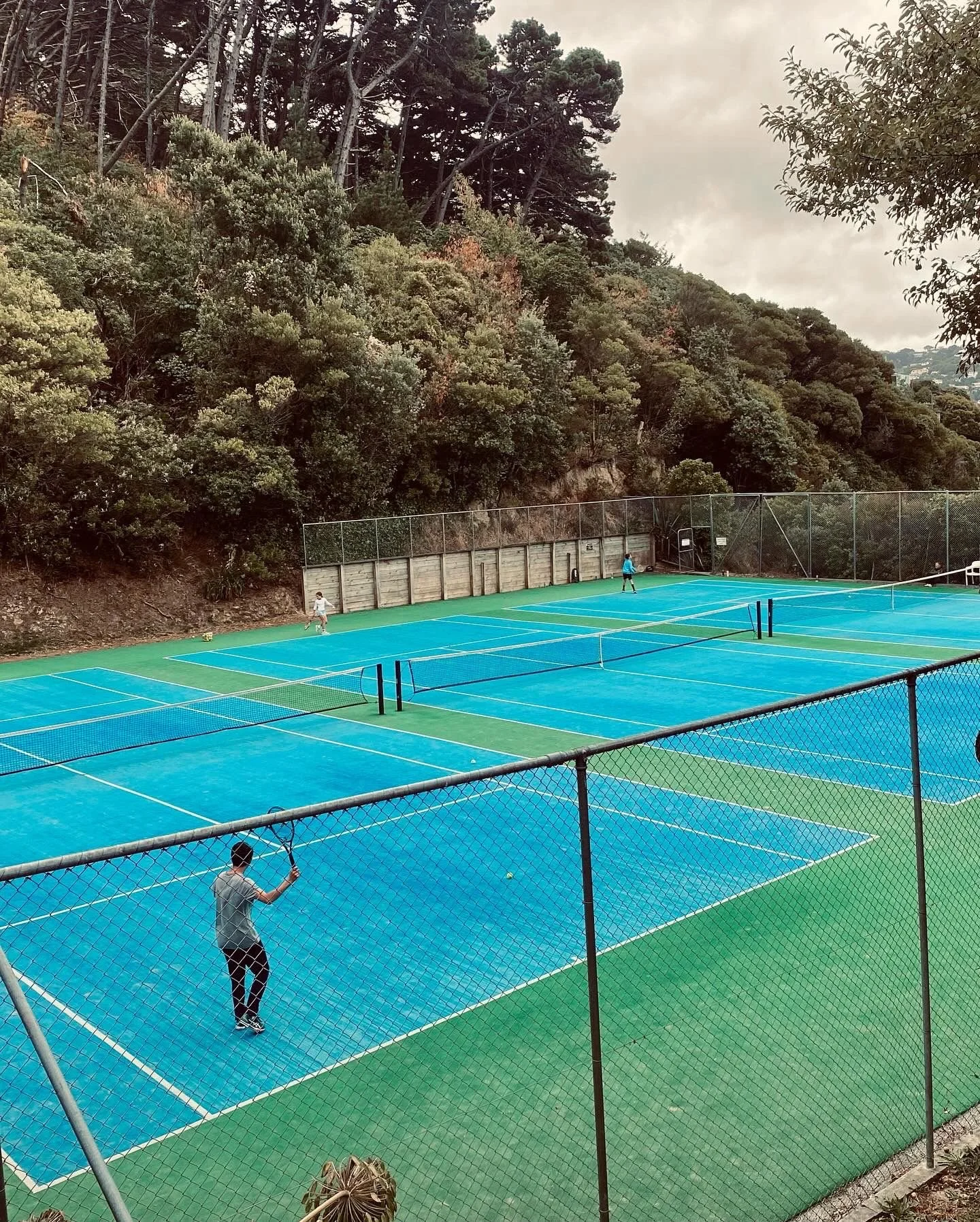 People playing tennis on multiple blue courts surrounded by a green fence and a wooded hillside.