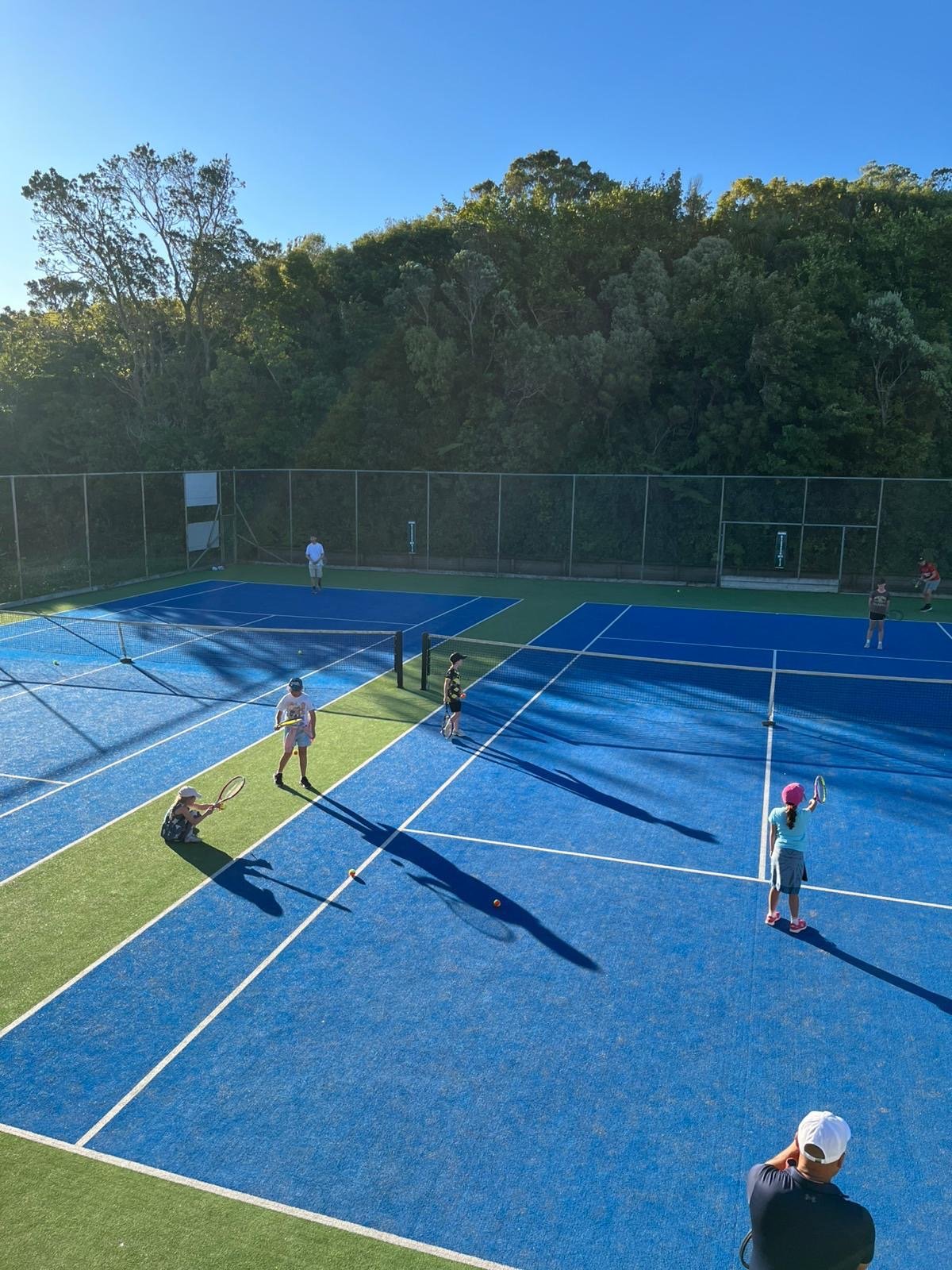 People playing tennis on outdoor courts with a wooded background under a clear blue sky.