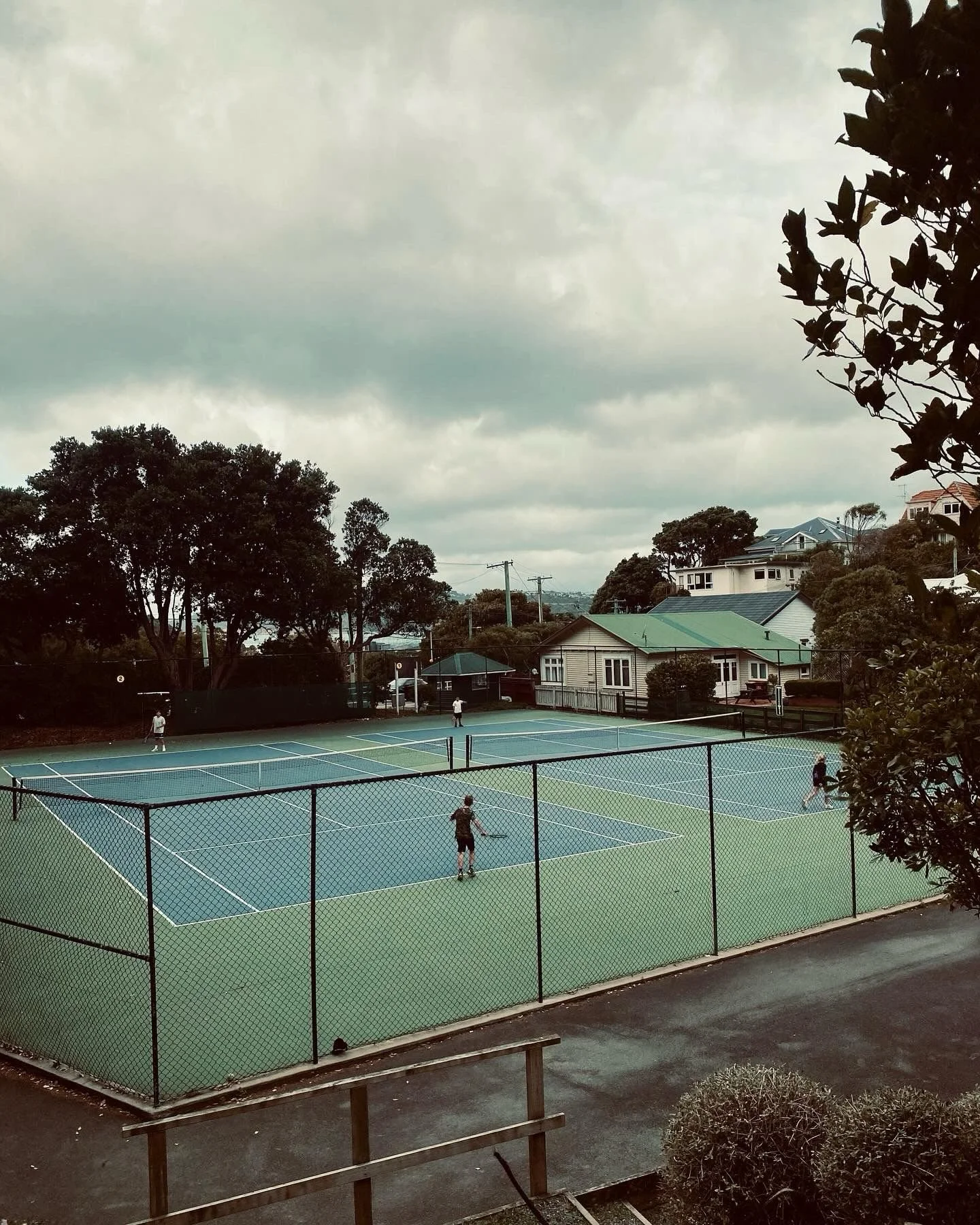 People playing tennis on outdoor courts surrounded by trees and residential houses under cloudy sky.