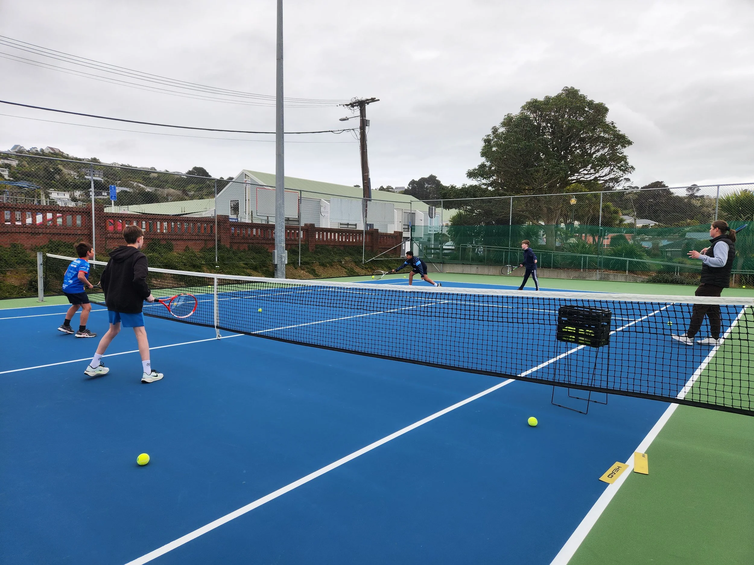 Children playing tennis on an outdoor court, with a coach watching, under overcast sky.