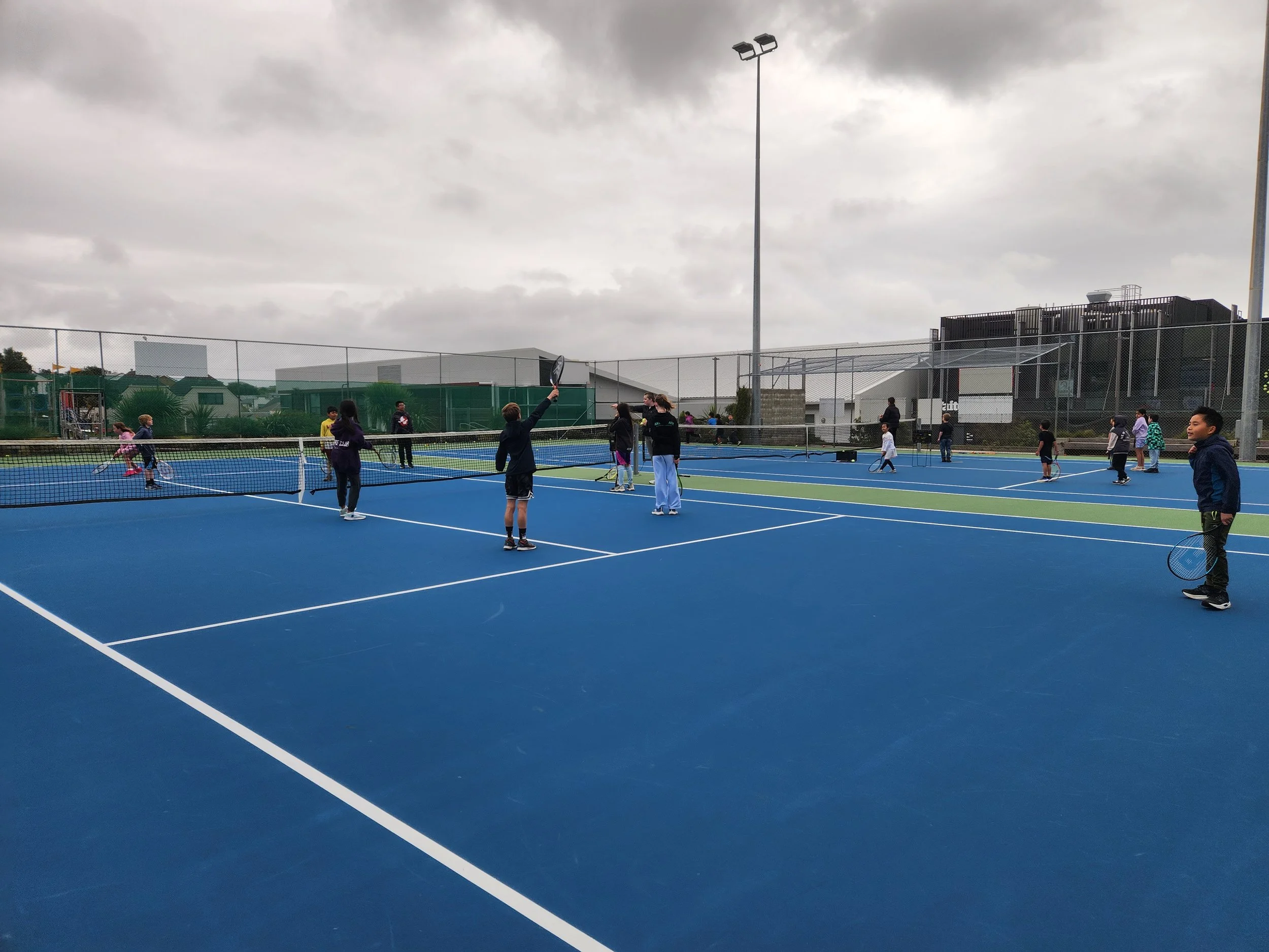Children and adults playing tennis on an outdoor blue tennis court under cloudy sky.