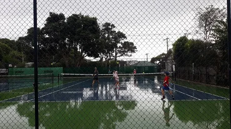 People playing tennis on a wet outdoor court with a chain-link fence, trees, and cloudy sky in the background.