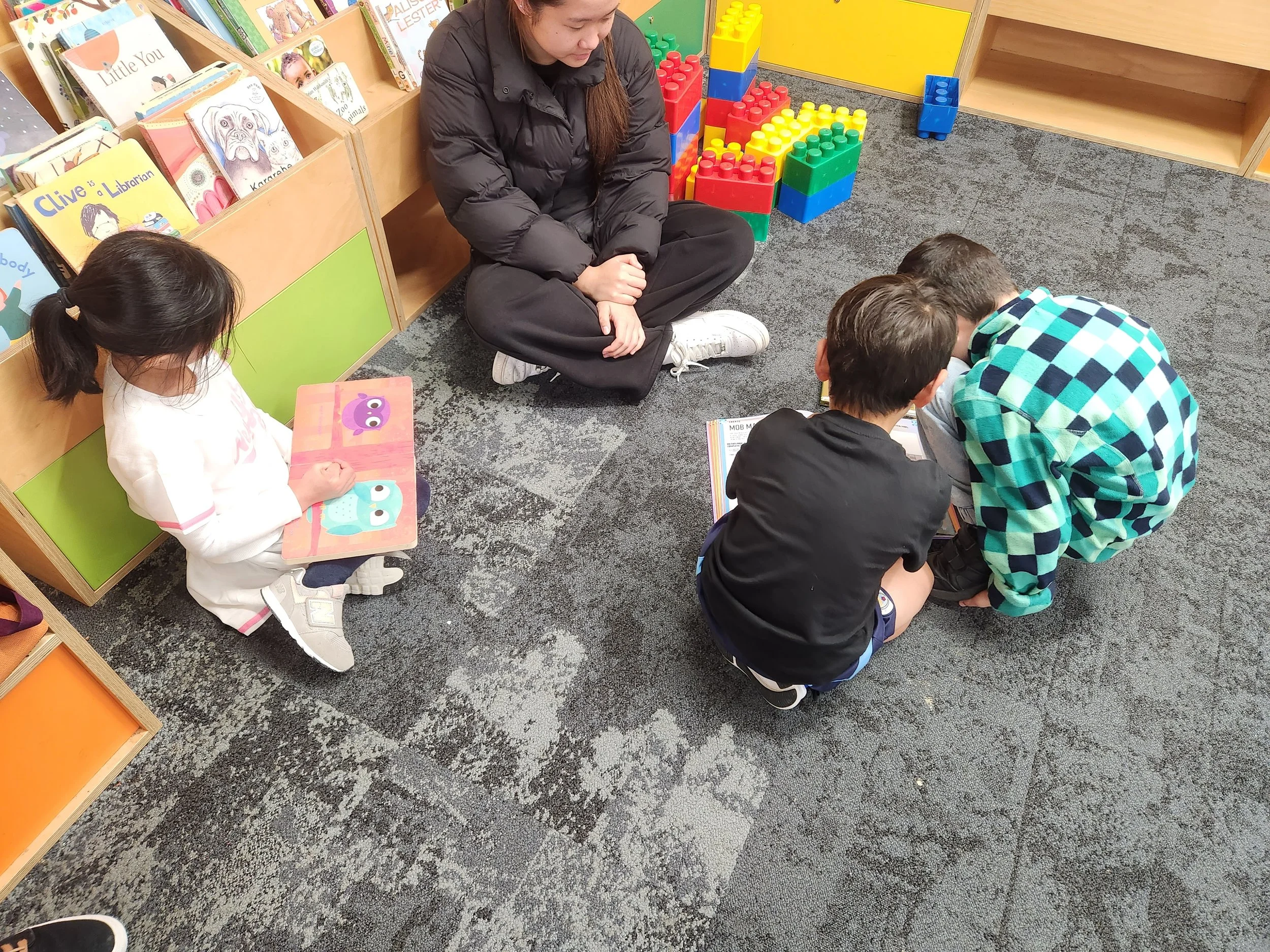 A woman and three children sitting on the carpet in a children's bookstore or library. The woman is sitting cross-legged, observing the children who are closely looking at a book on the floor. One girl, with a ponytail, is sitting alone on the left s