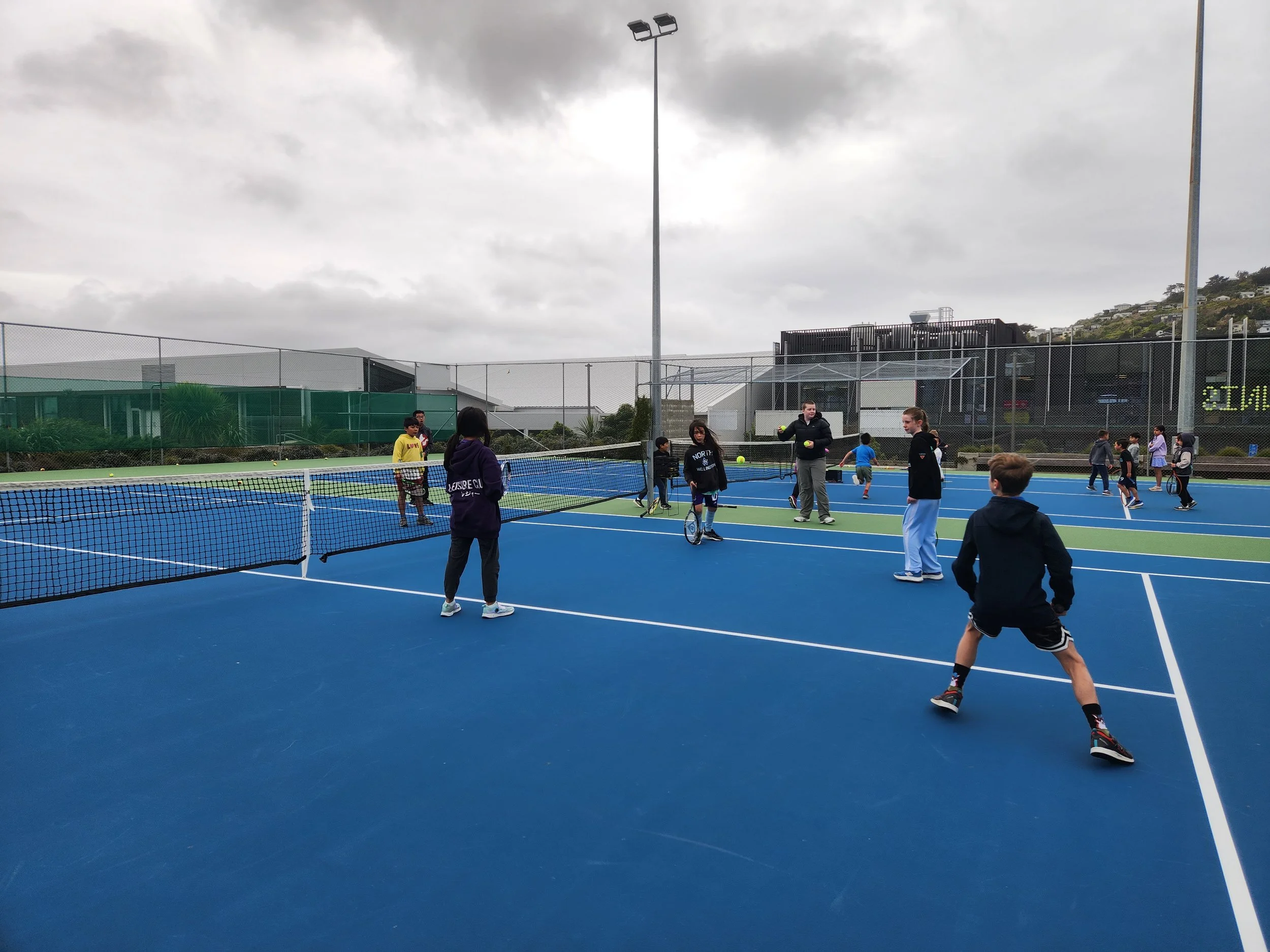 Kids and adults playing tennis on an outdoor blue tennis court with cloudy sky overhead.