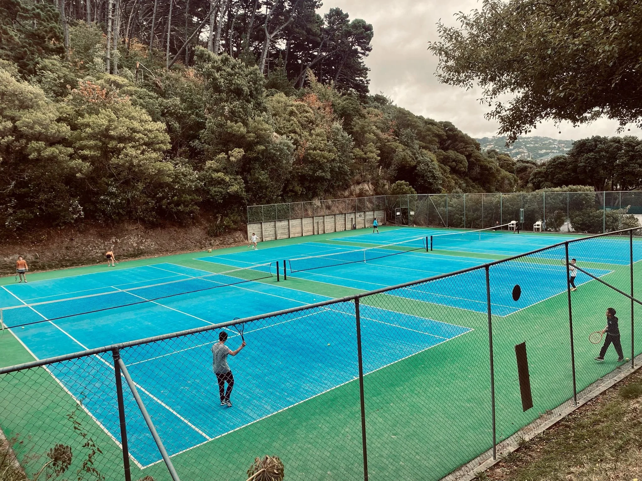 People playing tennis on multiple blue tennis courts surrounded by a chain-link fence and trees in the background.