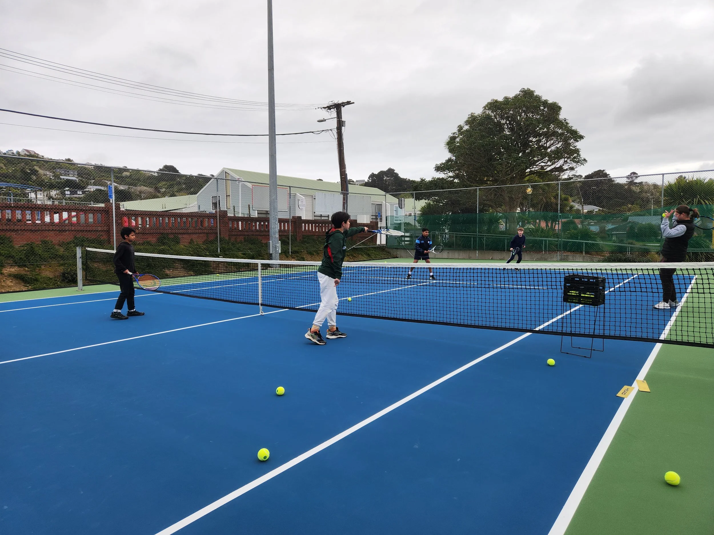 People playing tennis on an outdoor court with a blue surface, surrounded by a fence, under an overcast sky.