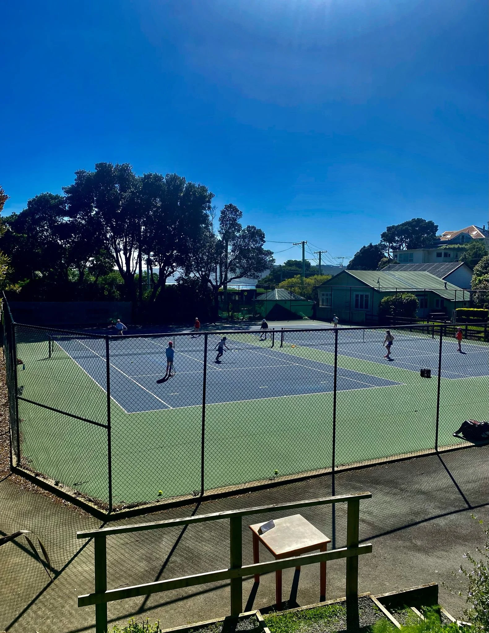 A group of children and adults playing tennis on an outdoor tennis court under a bright blue sky, surrounded by trees and residential houses.