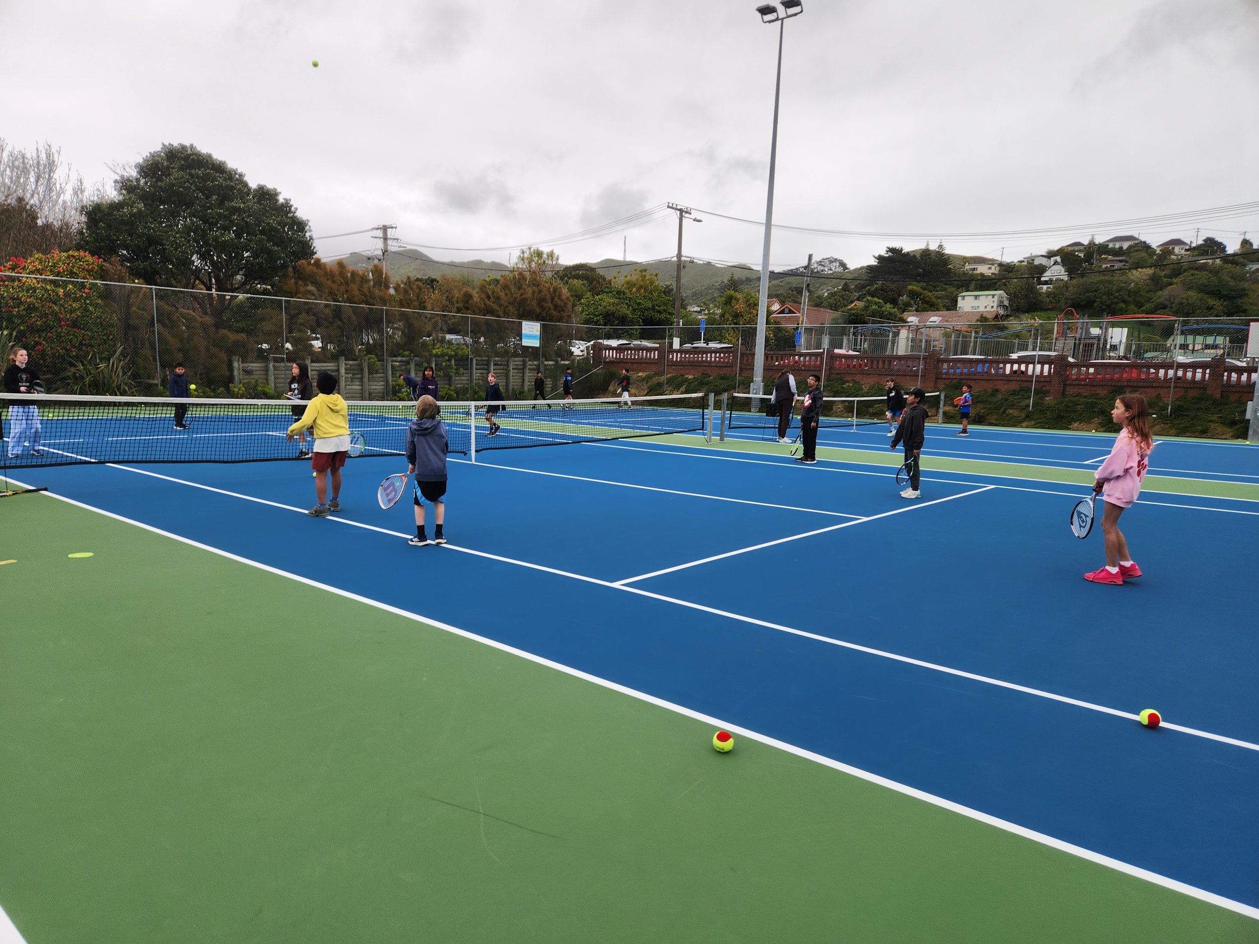 Children and teenagers playing tennis on an outdoor court during cloudy weather.