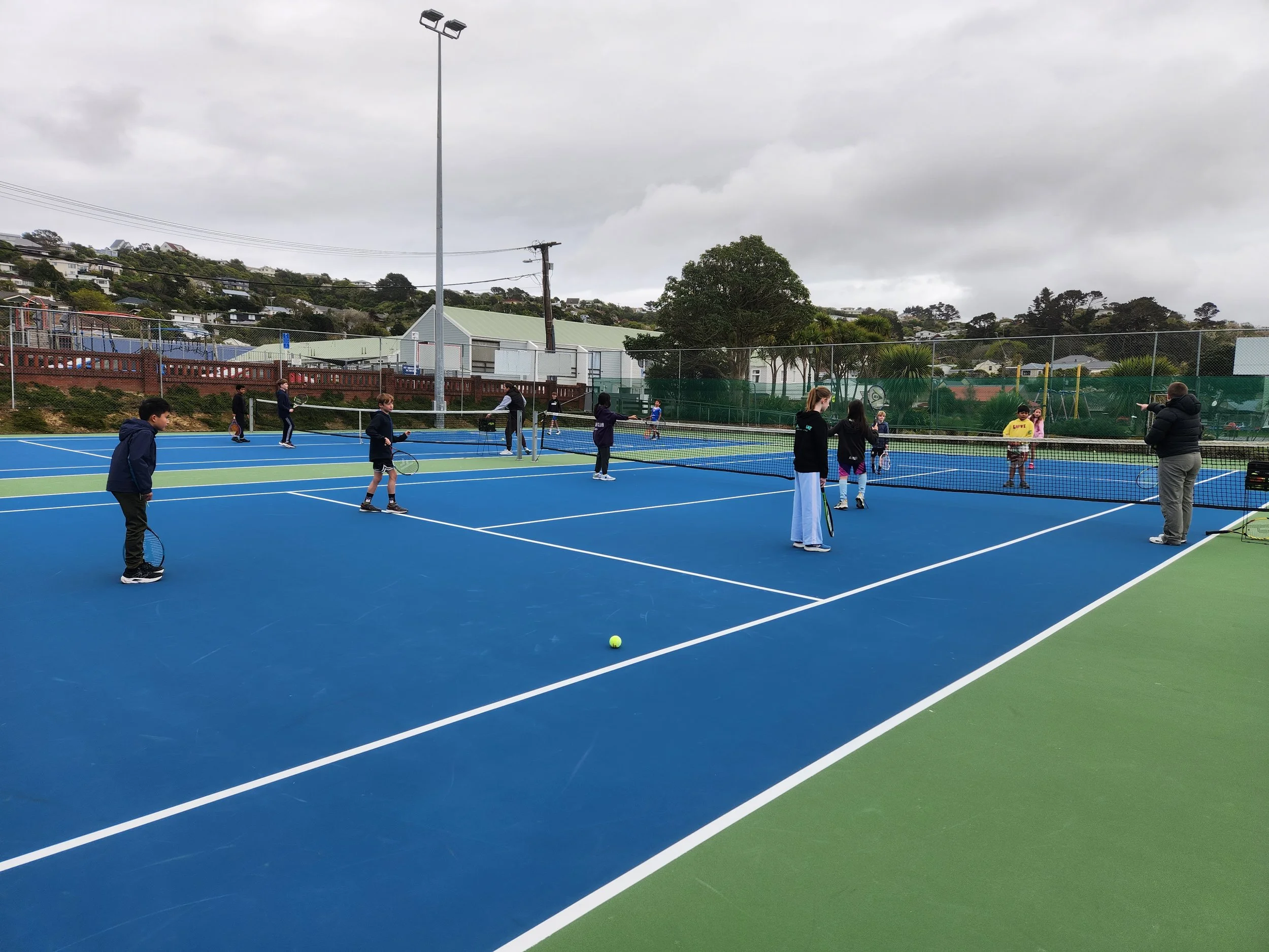 Children playing tennis on a blue and green outdoor tennis court, with a coach or instructor nearby and a cloudy sky overhead.