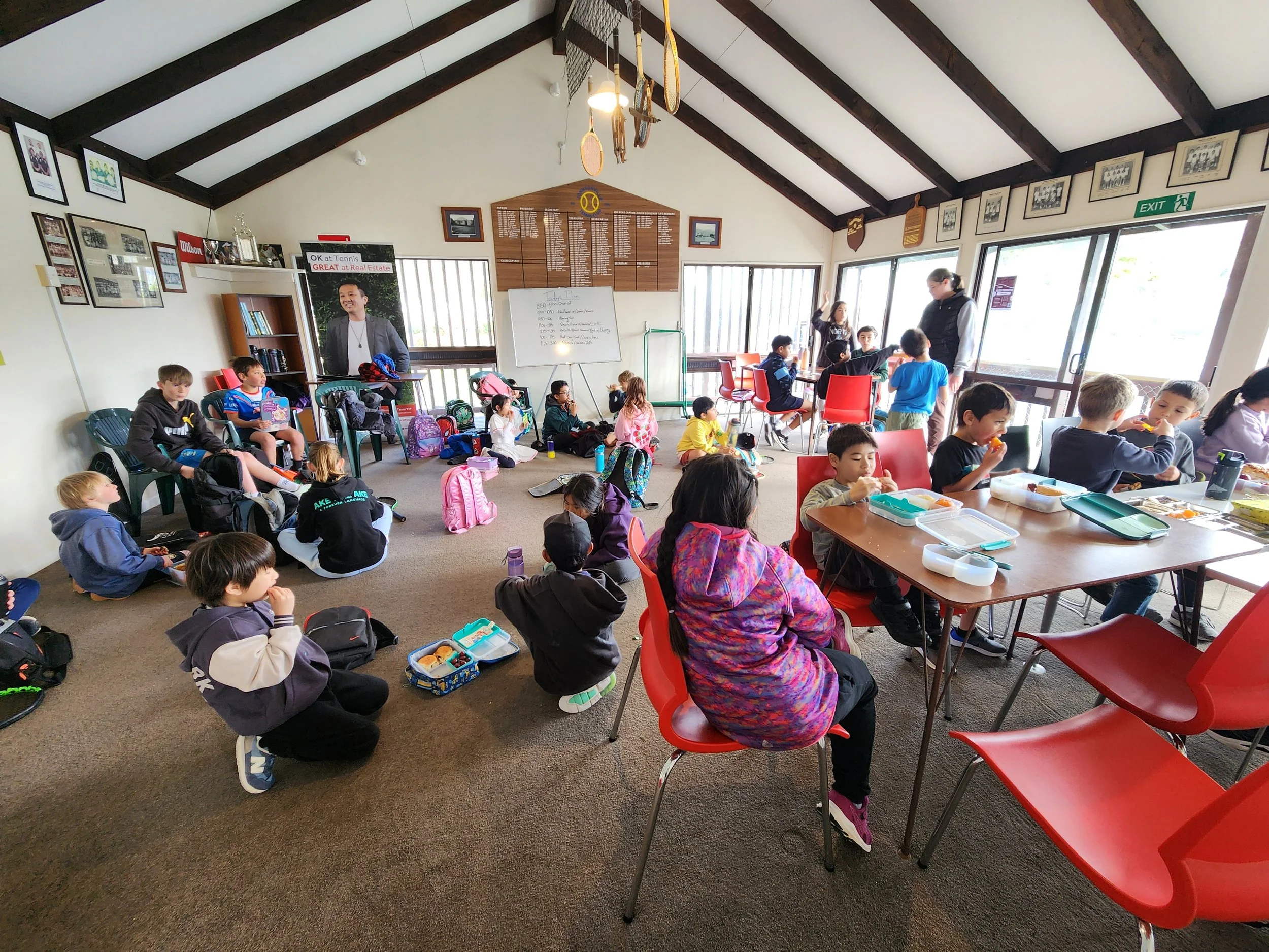 Children sitting on the floor and at tables in a classroom or community center, some eating lunch, with teachers or staff members standing near windows, and framed photos or awards on the wall.
