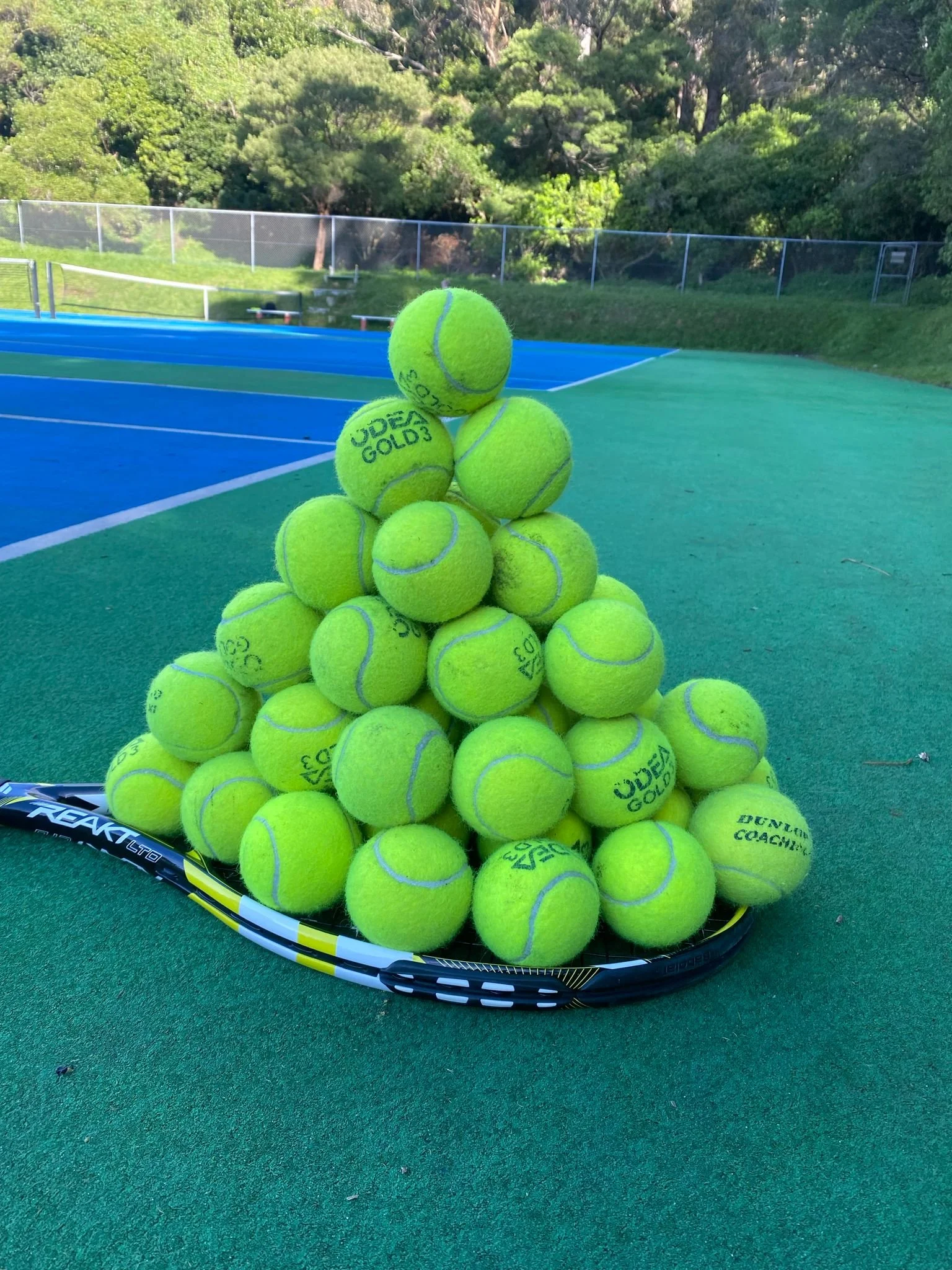 A pile of yellow tennis balls on a tennis racket on a green tennis court with trees and a fence in the background.
