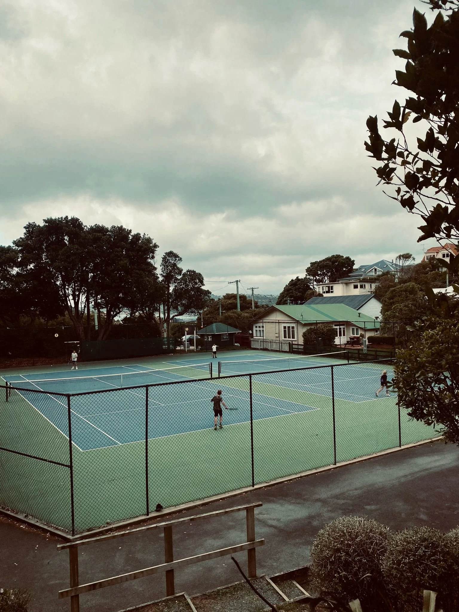 Five people playing tennis on an outdoor court with a green and blue surface, surrounded by a black chain-link fence, under a cloudy sky.