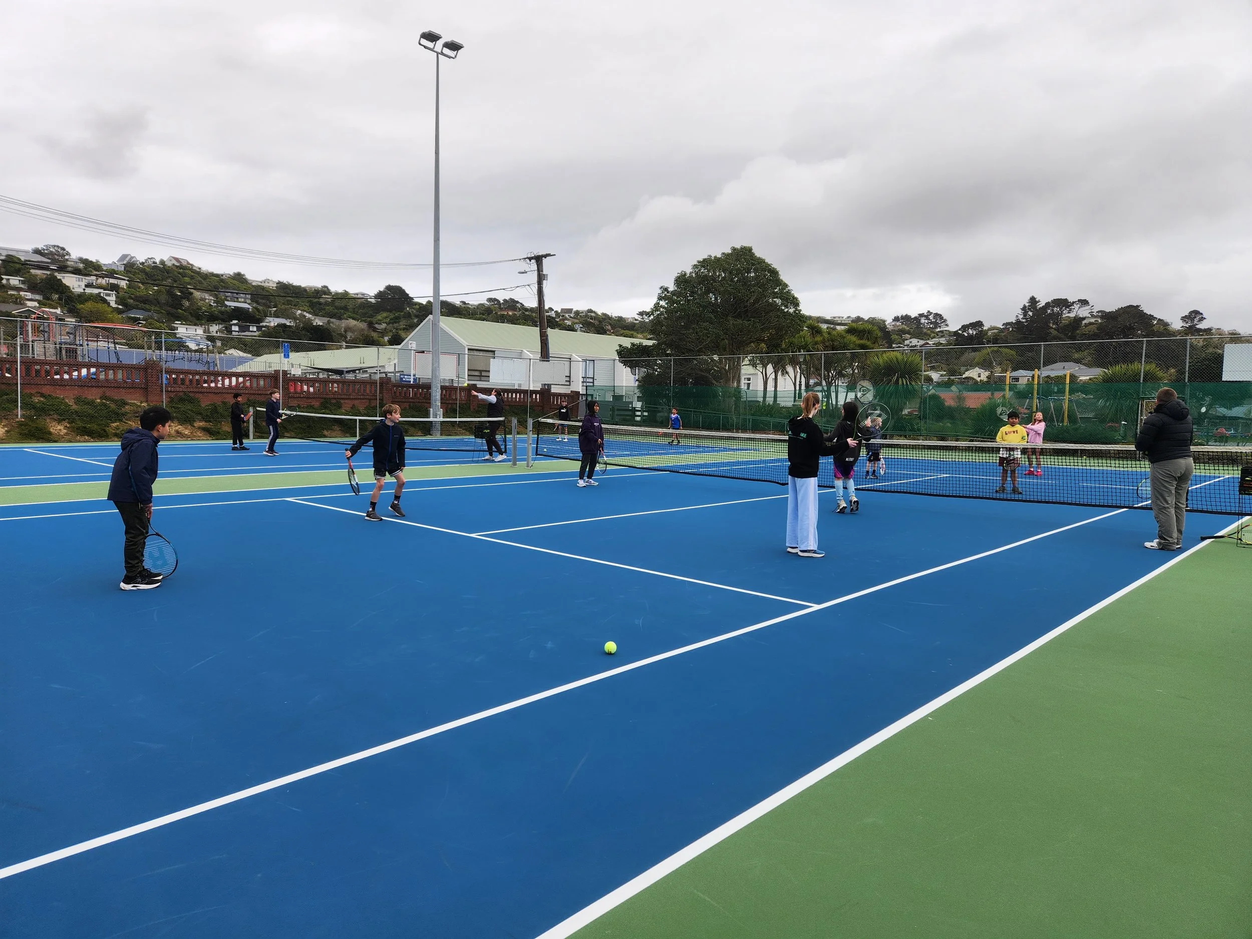 People playing tennis on an outdoor blue tennis court with a net, surrounded by a fence, with overcast skies.