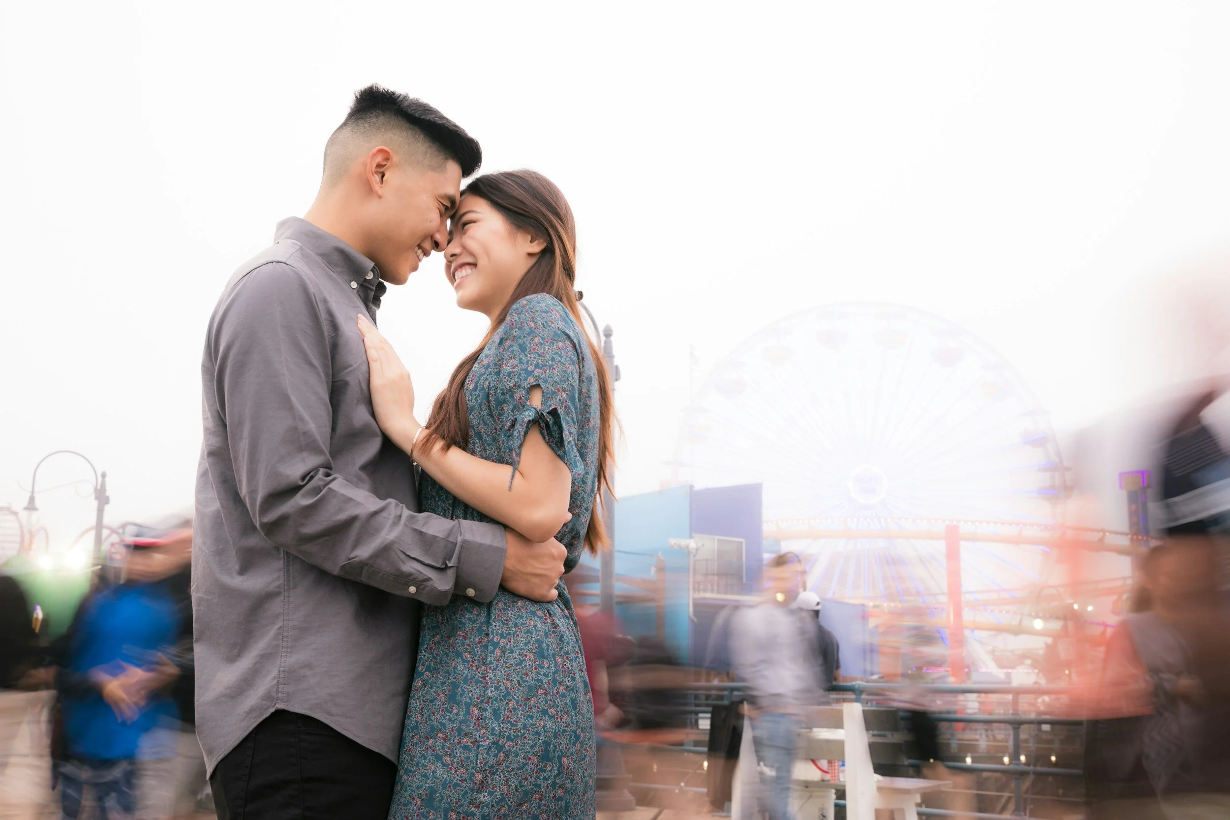 A young couple is smiling and touching foreheads at an amusement park with a Ferris wheel in the background, and several blurry people walking around.