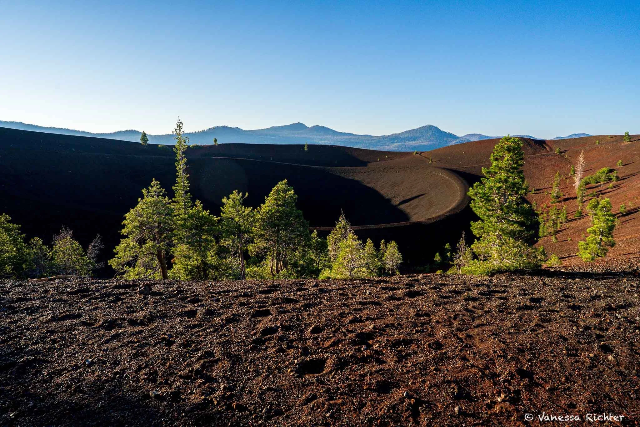 Looking down from the upper rim of Cinder Cone in Lassen Volcanic National Park to the lower rim trail circling the crater, with pines below.