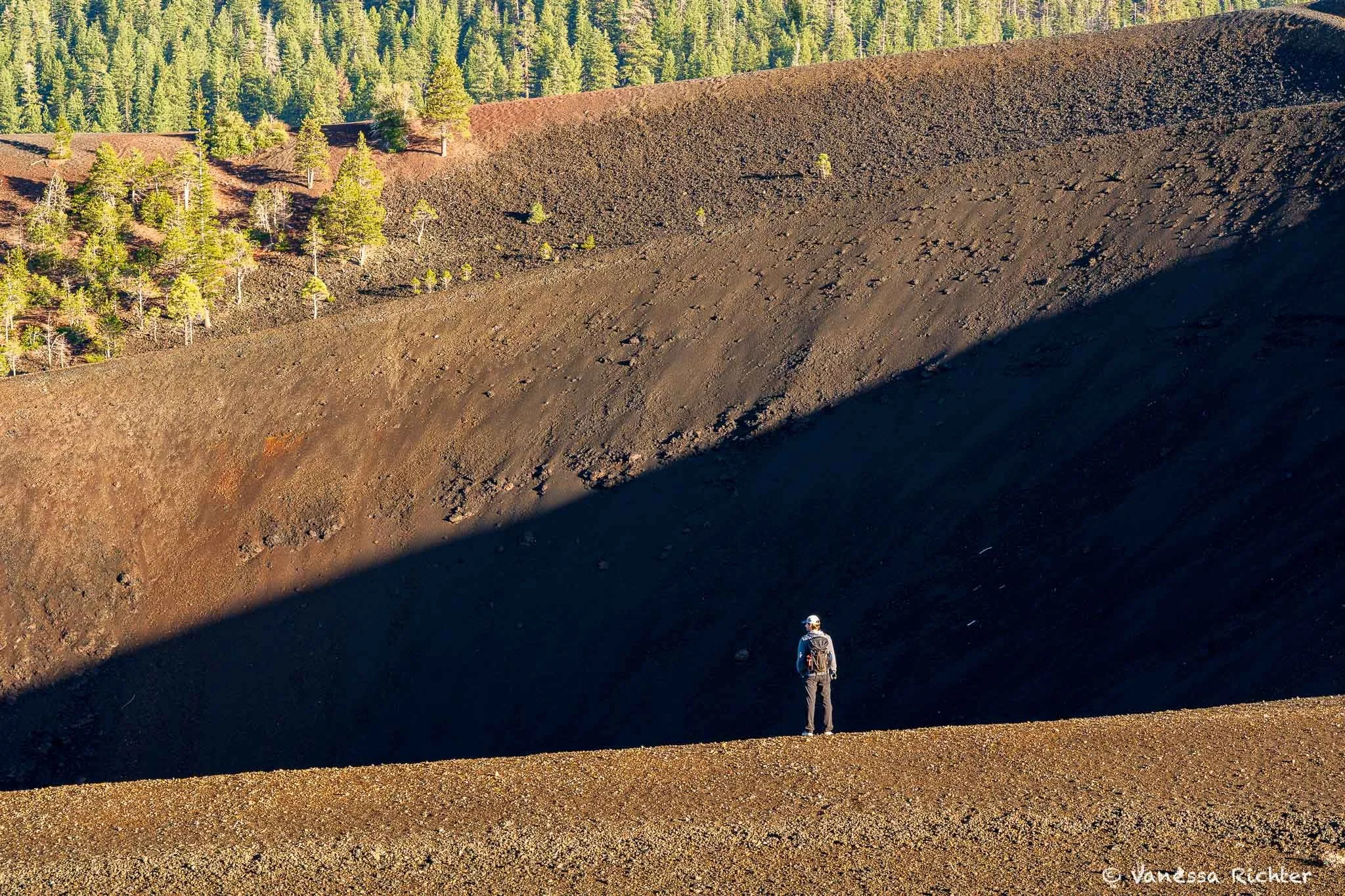 iker standing on the rim of Cinder Cone in Lassen Volcanic National Park, a large shadow points into the crater with pine forest in the distance.