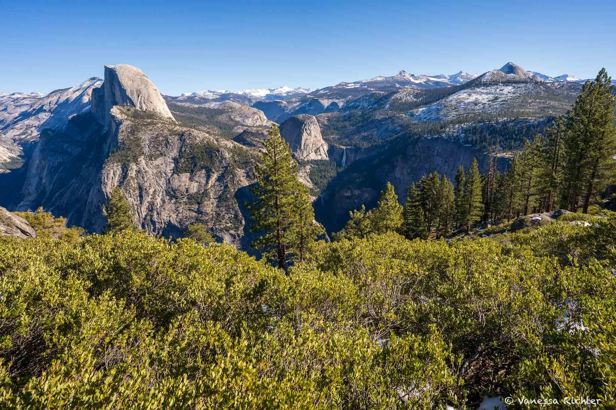 Glacier Point view in Yosemite National Park with Half Dome on the left side of the picture, and Vernal and Nevada Falls in the center. The tallest mountain peaks are dusted in snow.