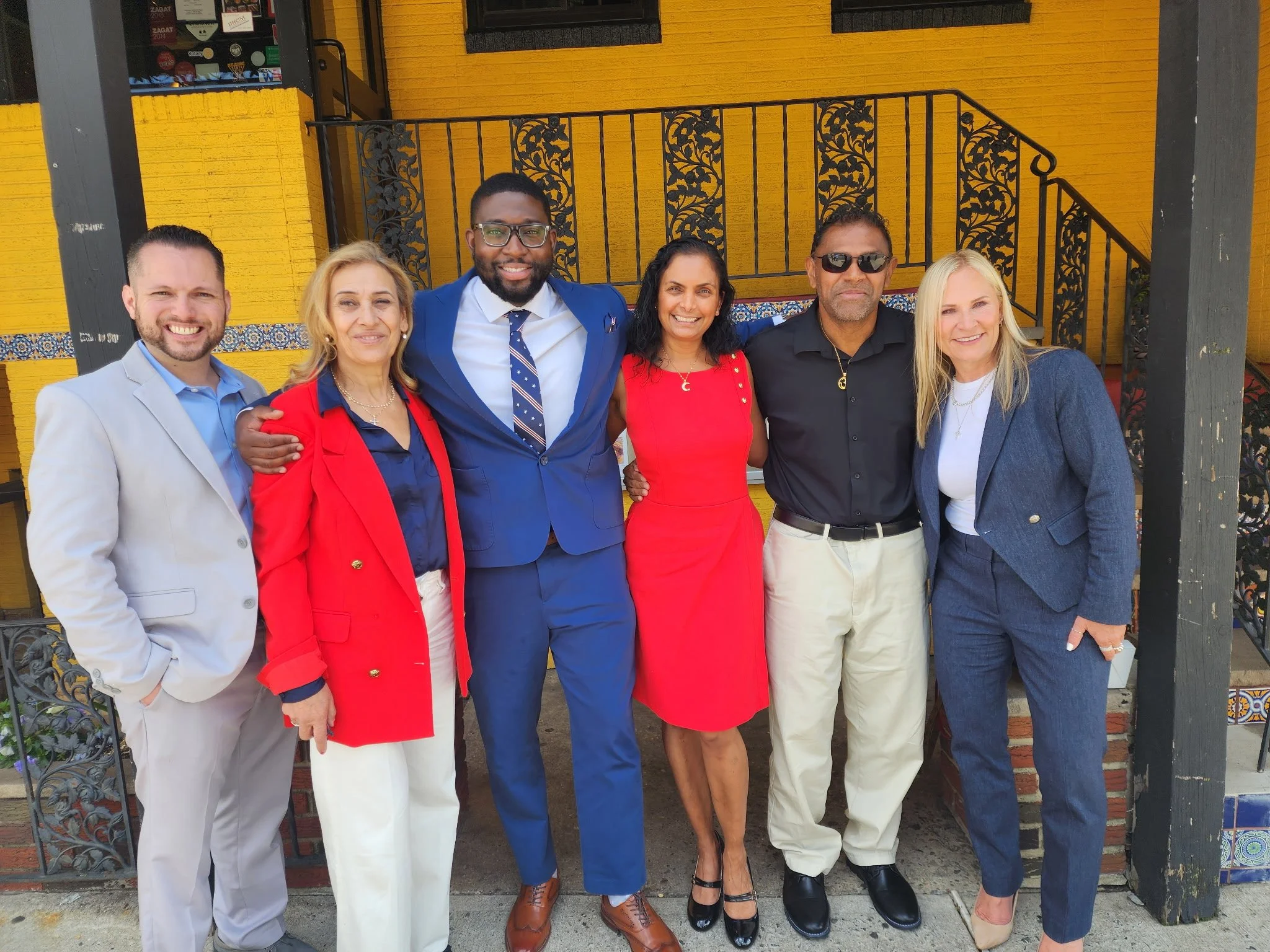 Group of six people standing together in front of a yellow wall with black wrought iron details, dressed in business or semi-formal attire, smiling at the camera.