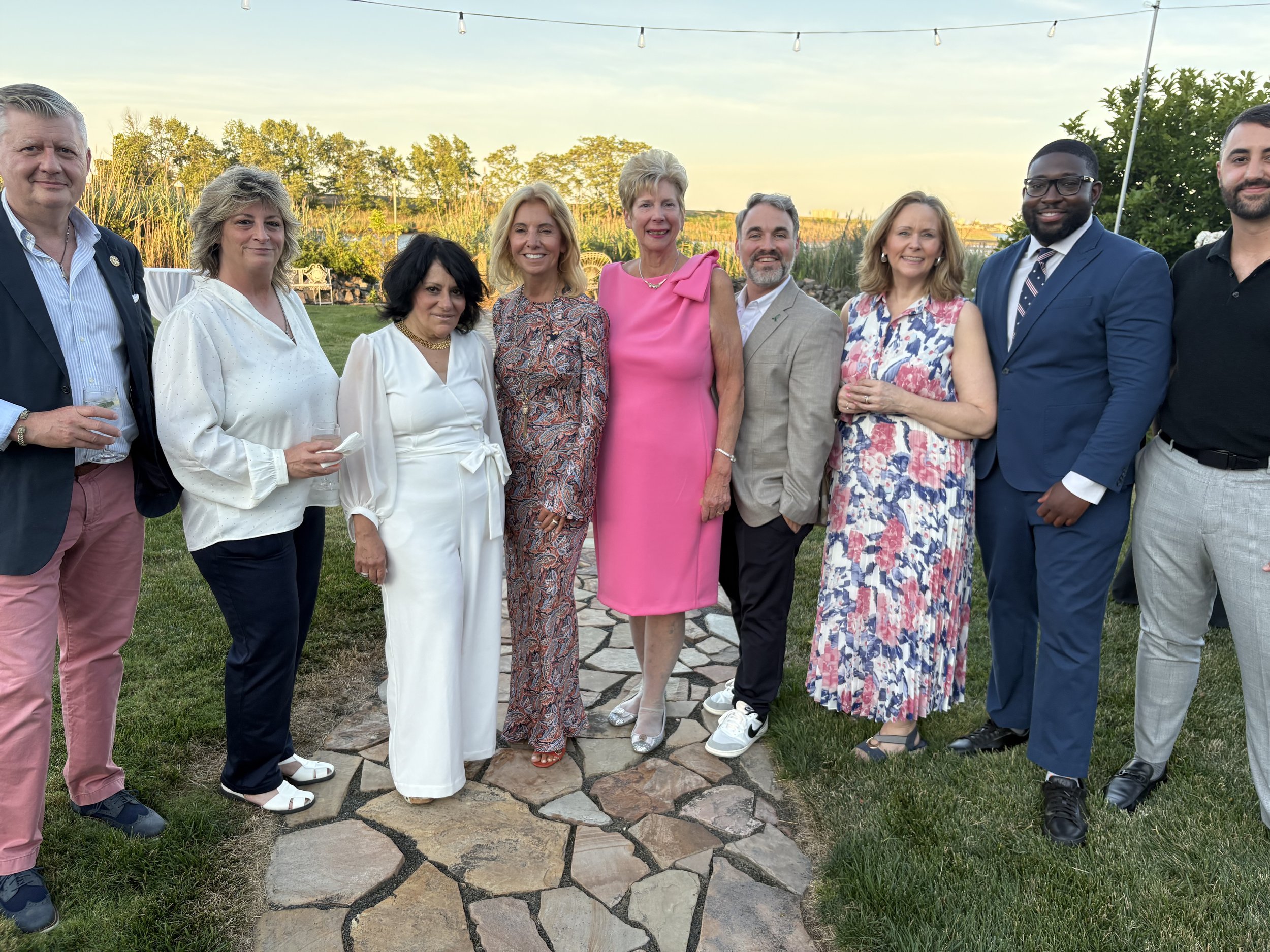 A group of ten people standing outdoors on a stone pathway, smiling at the camera, with trees and a sunset sky in the background.