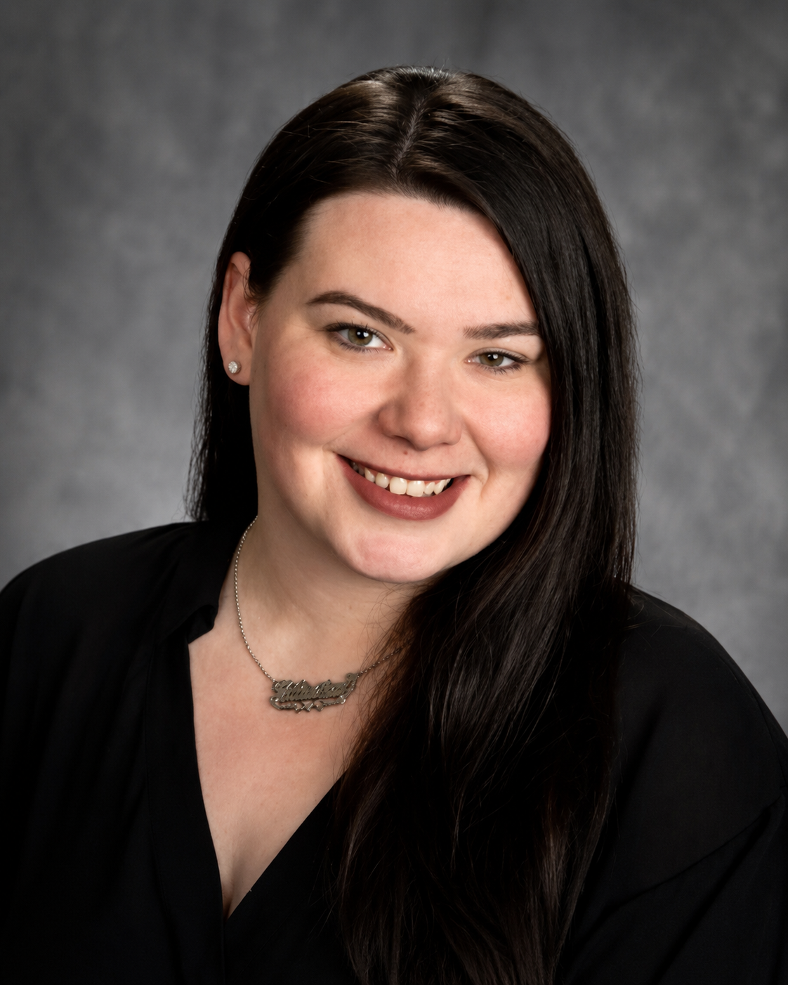 Head and shoulders portrait of a smiling woman with dark brown hair, wearing a black top, earrings, and a necklace with a name pendant, against a gray background.
