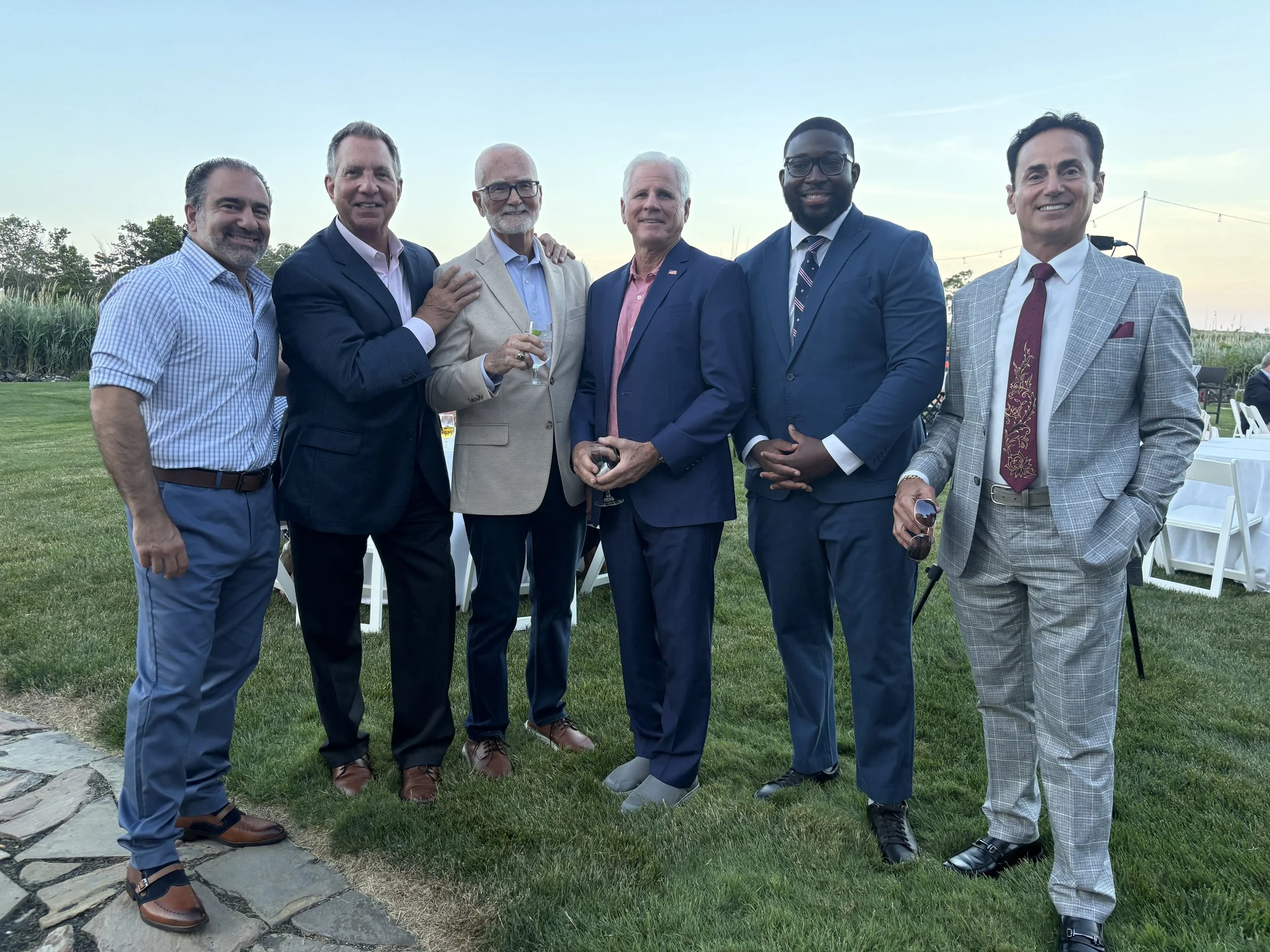 A group of six men in suits and business casual attire standing together outdoors on a grassy area during early evening, smiling at the camera.