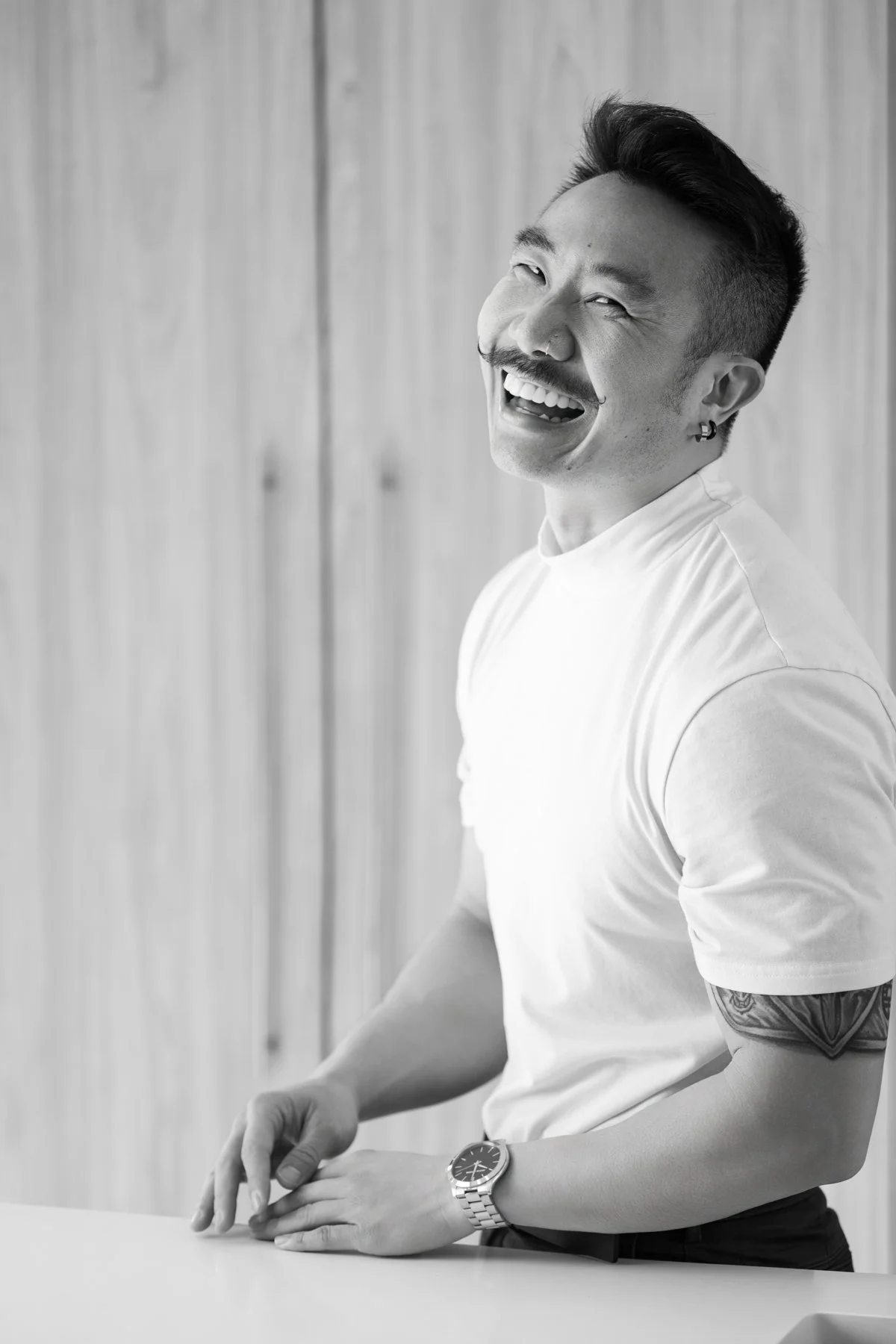 Black and white photo of a man with short, styled hair, a mustache, and earrings, smiling and laughing. He is wearing a white t-shirt, a watch, and has a tattoo on his forearm. He is sitting at a table with a wooden wall background.