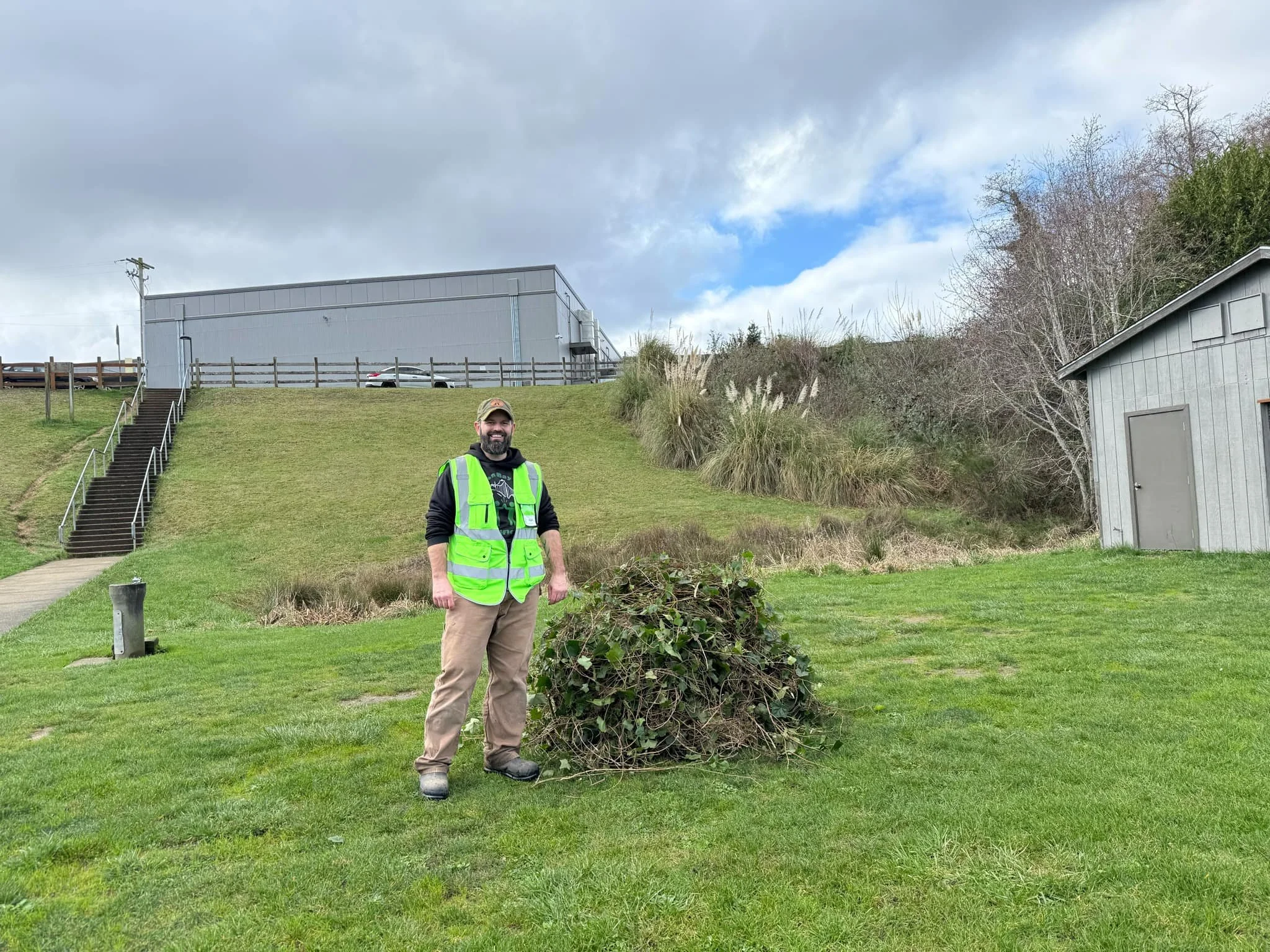 an OBAB volunteer stands next to a pile of cut English Ivy removed from the bleachers and the trails.