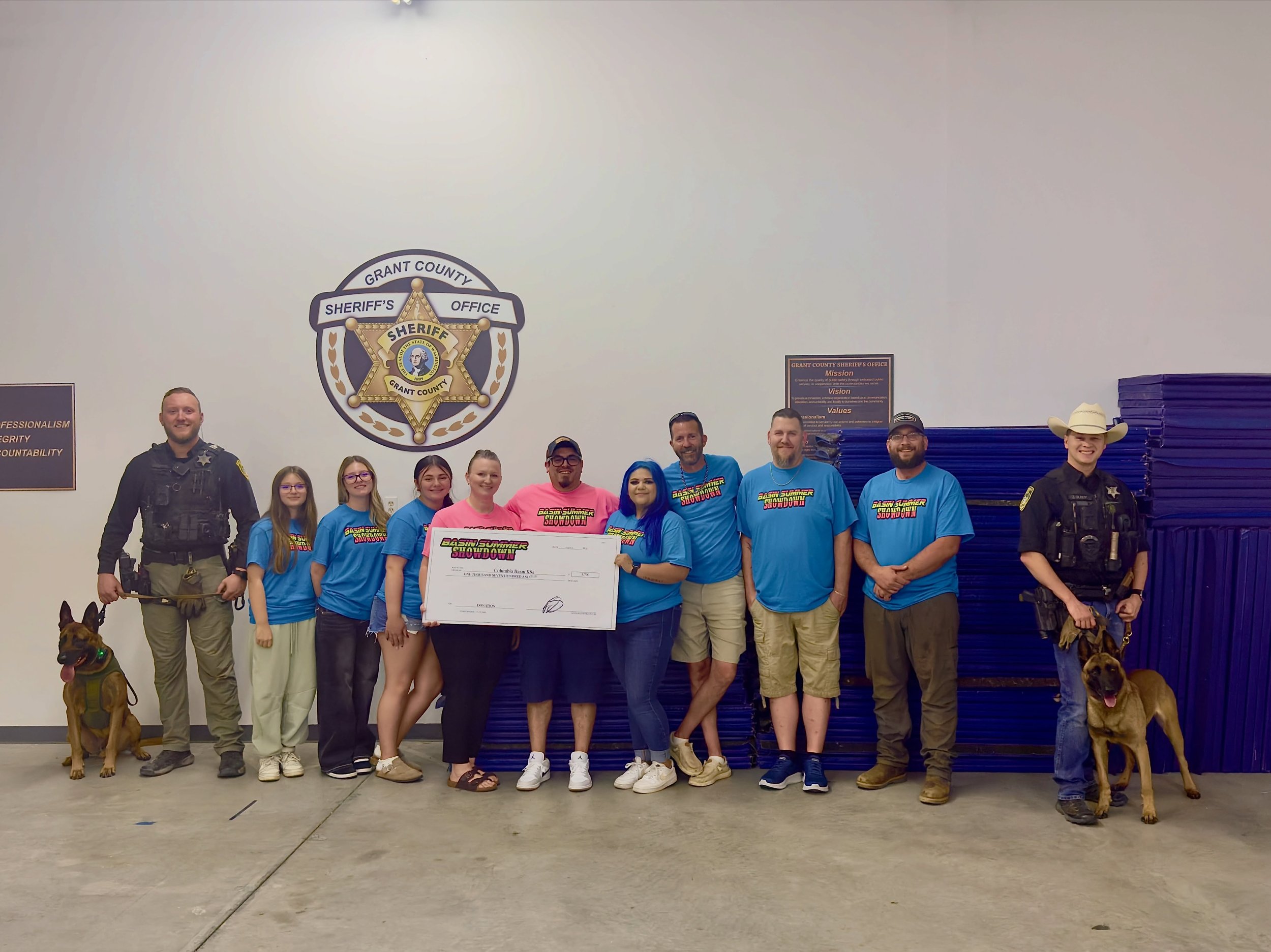Group of people, including children, adults, and police officers, standing together inside a building with a large sheriff's badge on the wall behind them. Some people hold a large check, and two dogs are present, one sitting and one standing with a police officer. The setting suggests a community or awards event at a sheriff's office.