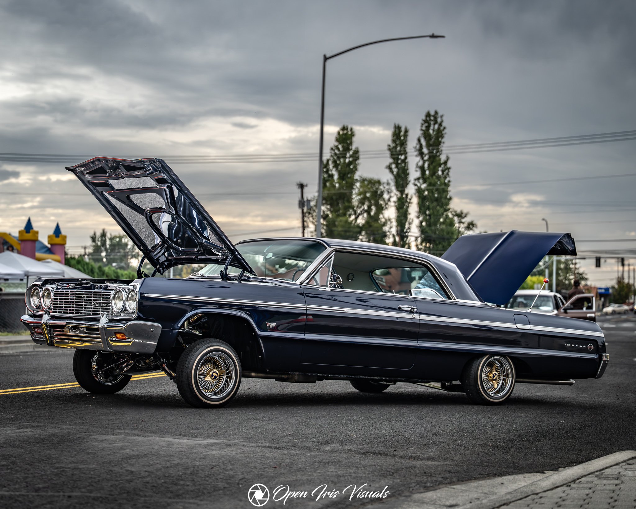 A classic black Chevrolet Impala with gold accents and custom wheels is parked outdoors on a cloudy day, with its hood open revealing the engine. In the background, there are trees, a streetlight, power lines, and a colorful inflatable play structure