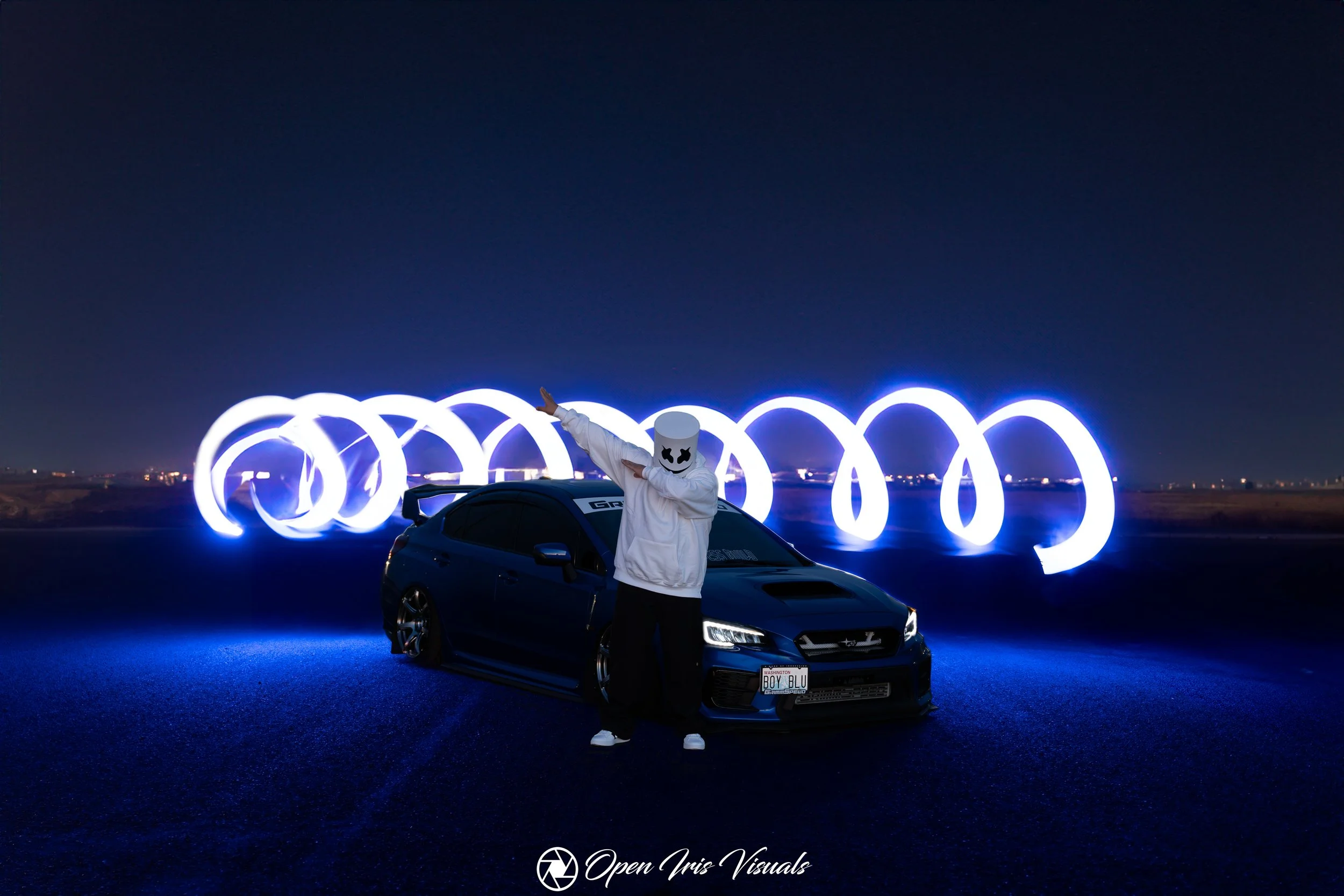 Person wearing a marshmallow mask posing in front of a black sports car on a dark open road at night, with blue LED light trails in the background.