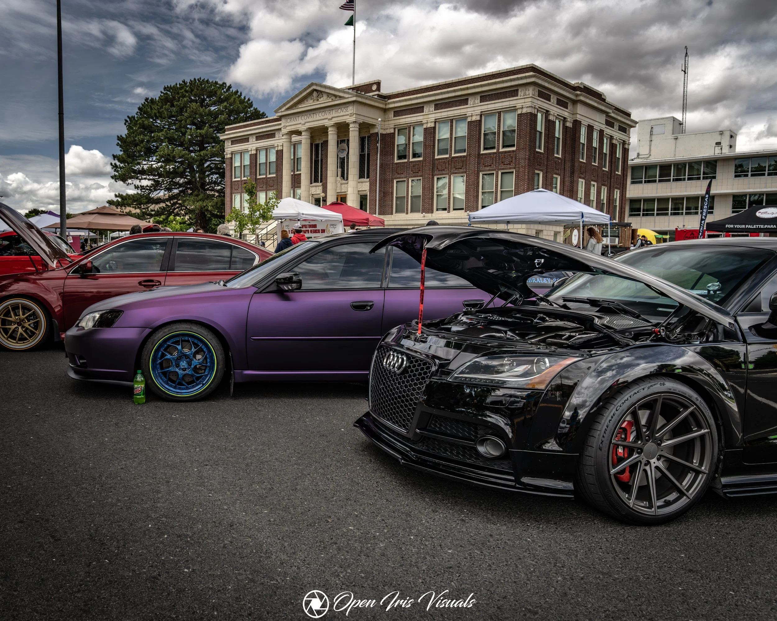 A lineup of modified cars at a car show in front of a historic courthouse building with tents and people around.