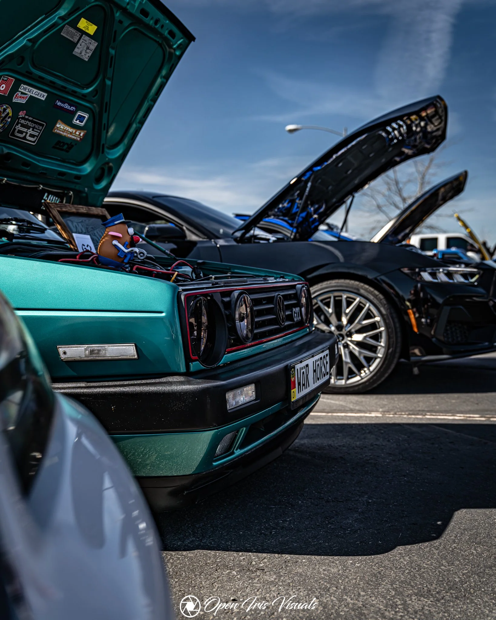 Close-up of a teal vintage  VW car with its hood open at a car show, displaying stickers on the inside of the hood and a small plush toy on the engine. Several modern black cars with their hoods open are in the background, under a partly cloudy sky.