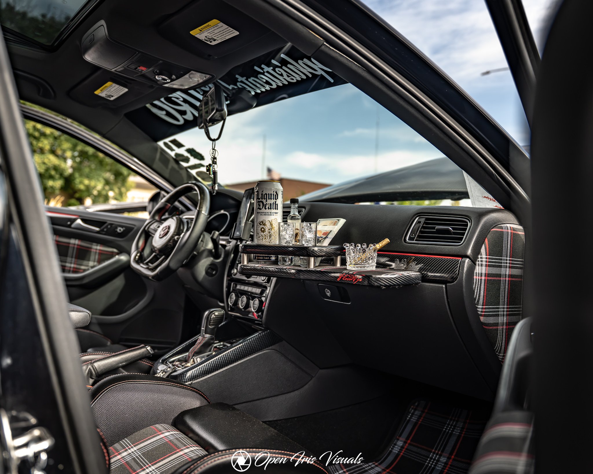 The interior of a car with a plaid-patterned seat, a bottle of Liquid Death water, glasses, and a small tray in the dashboard area; seen through the windshield with a blue sky and trees outside.