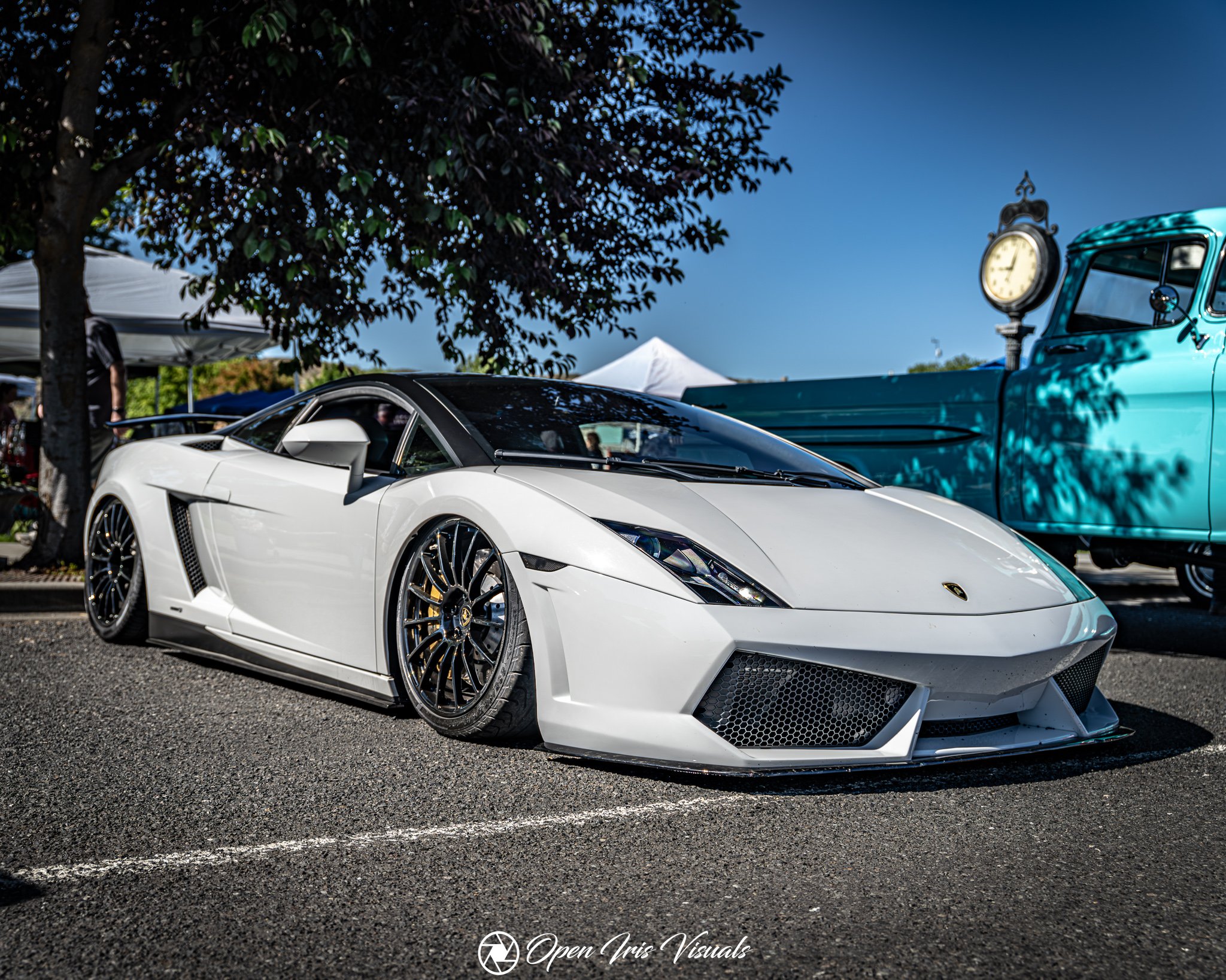 A white luxury sports car, likely a Lamborghini, parked on a street with a black roof, black wheels, and sleek design, next to a vintage teal truck with a clock mounted on its side. There is a tree with dark foliage casting shadows, and tents and peo