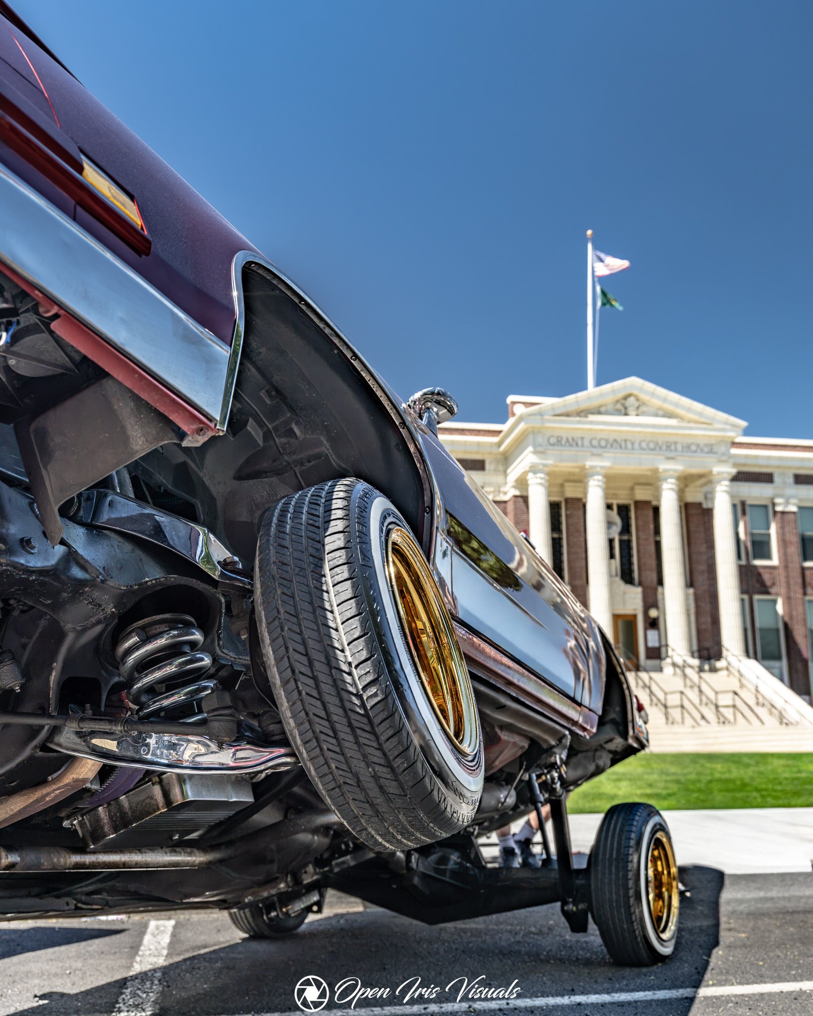 A close-up side view of a lowered black luxury car with gold rims on a trailer, parked in front of a city hall with the American and another flag flying on a clear sunny day.