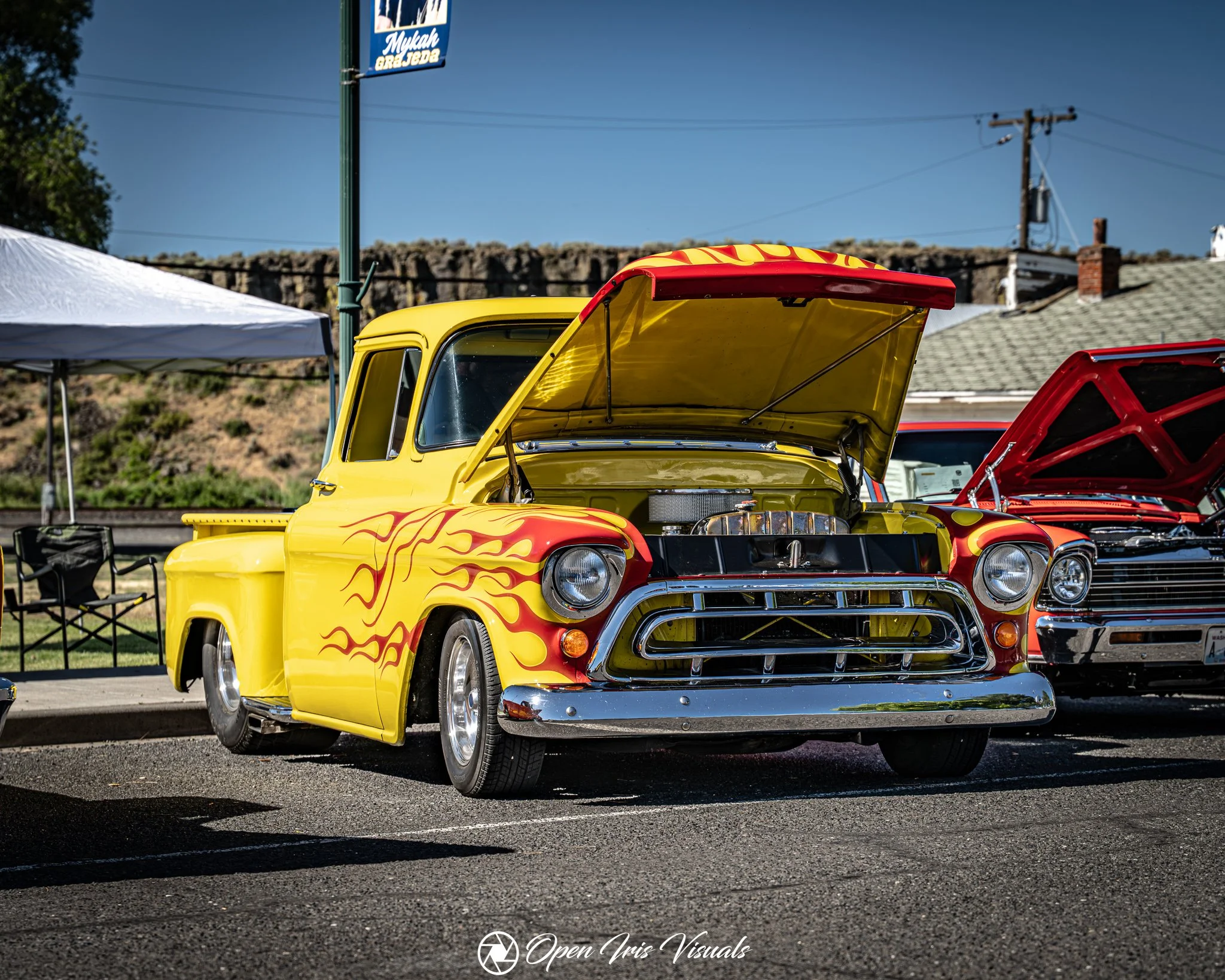 A vintage yellow pickup truck with orange flame decals on the sides, displaying its engine with the hood open at a car show. A white tent, a sign, and utility poles are visible in the background under a clear blue sky.