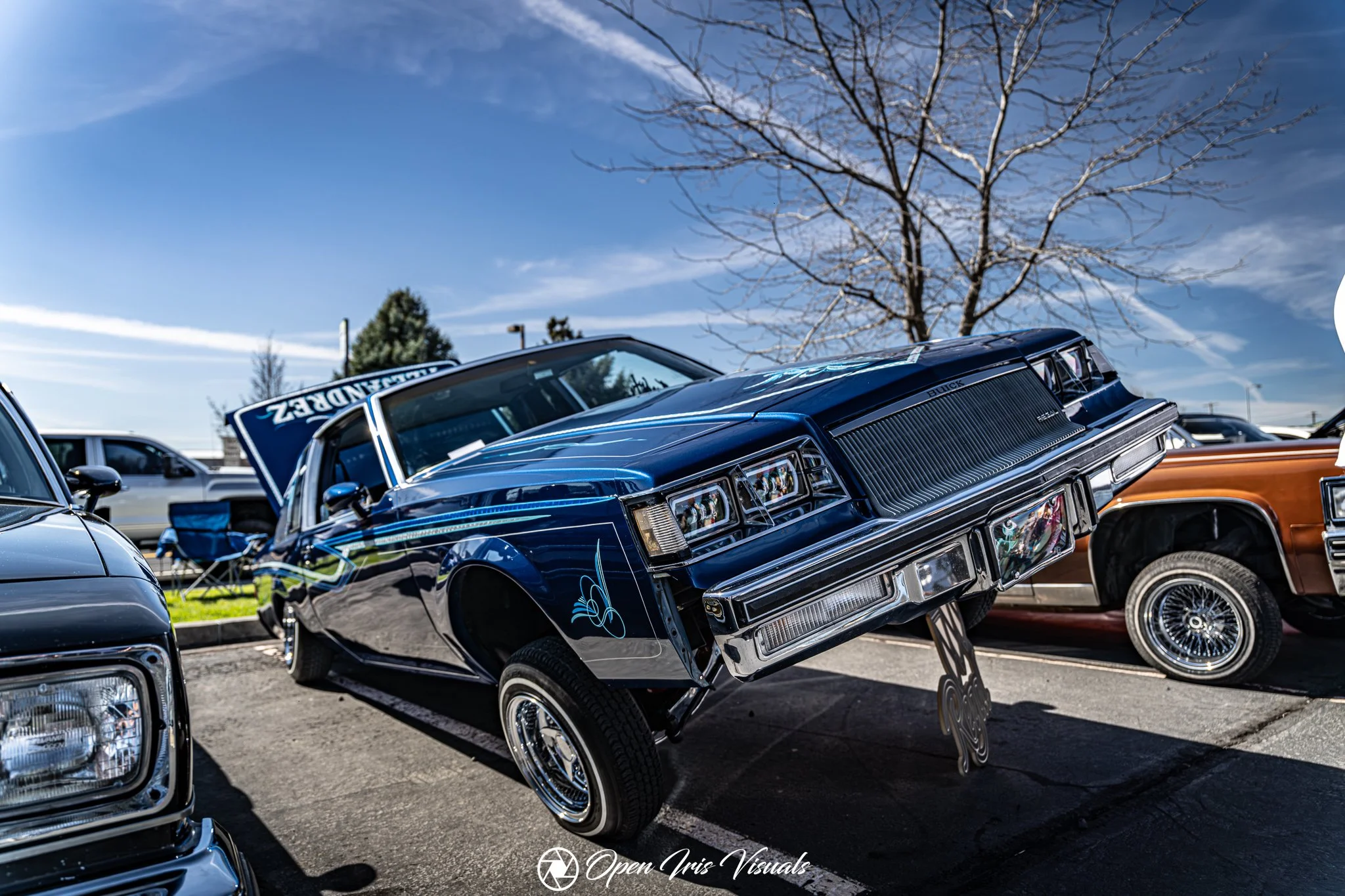 Blue vintage Buick lowrider on display at a car show, with its front wheels lifted off the ground, surrounded by other classic vehicles.