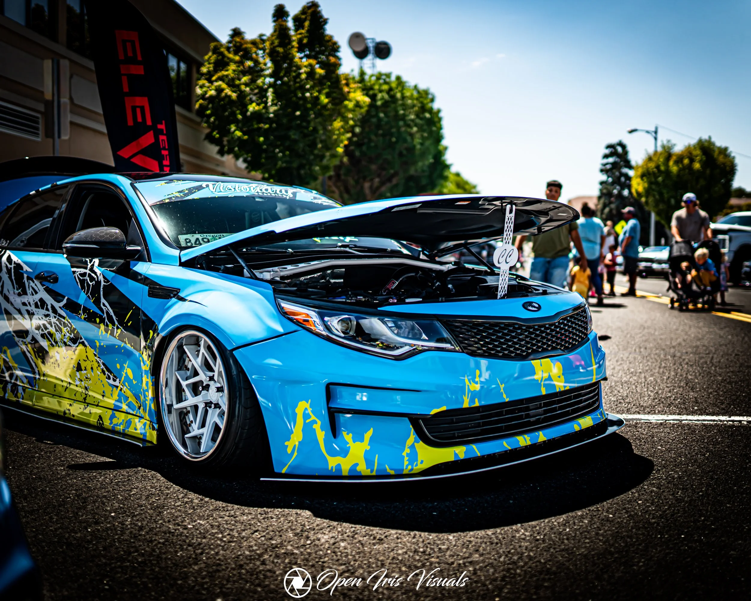 Blue sports car with custom paint and open hood at an outdoor car event, with people in the background.