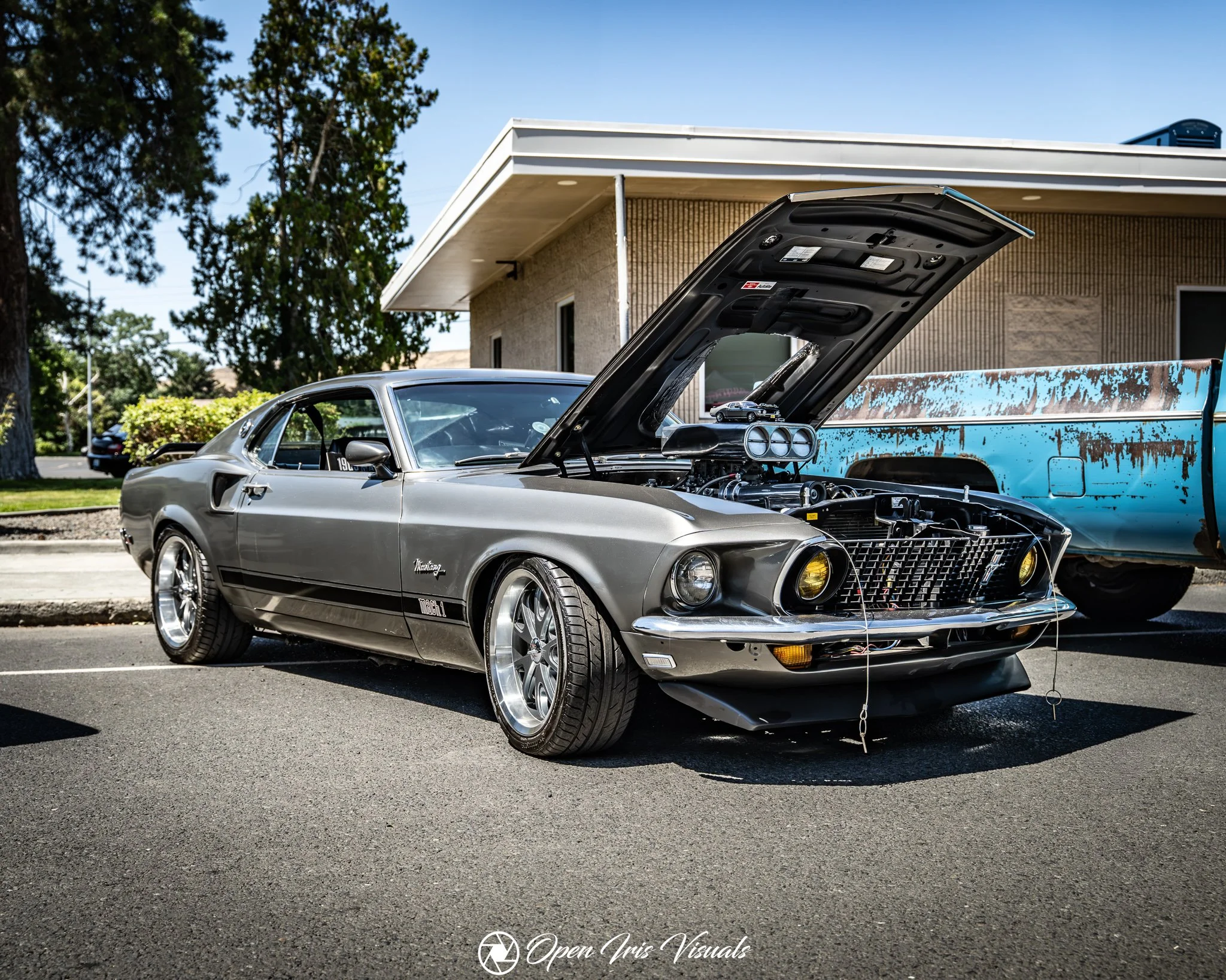 A classic gray Ford Mustang has its hood open, revealing the engine, parked in a parking lot near a beige brick building with a flat roof. A small model car is placed inside the engine bay. Trees and a clear blue sky are in the background.